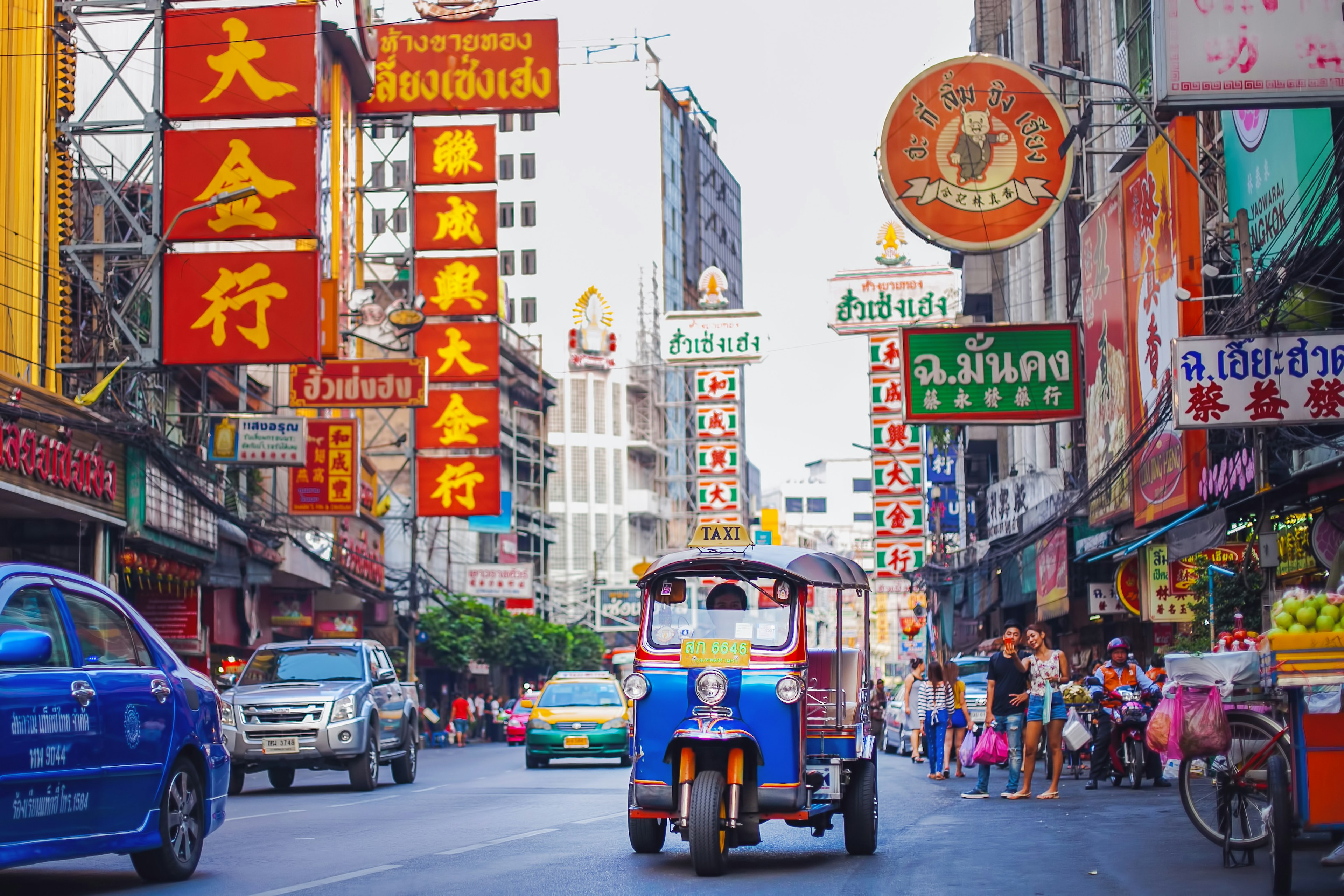 Cars and small motorized vehicles (tuk-tuks) drive down a busy street lined with neon signs