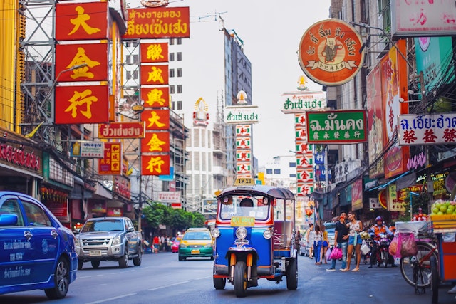 Cars and small motorized vehicles (tuk-tuks) drive down a busy street lined with neon signs