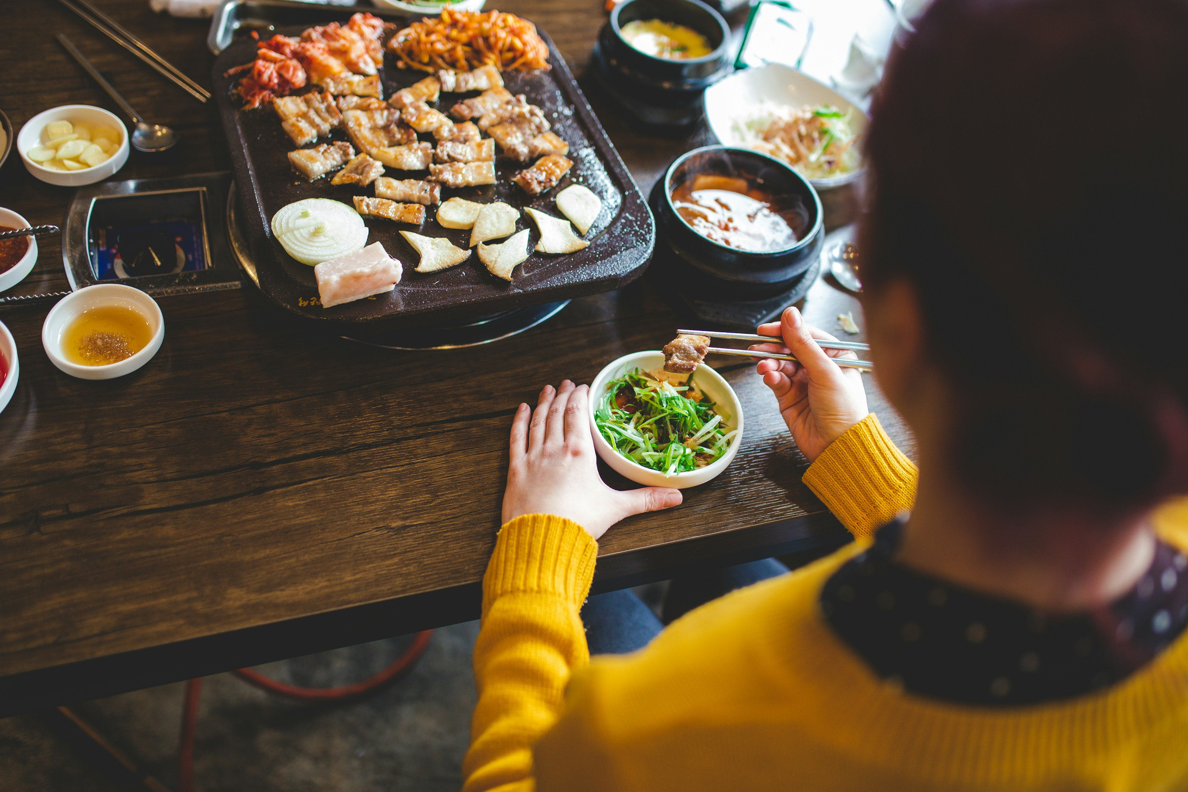 Rear view of a woman using chopsticks to eat a spread of Korean food