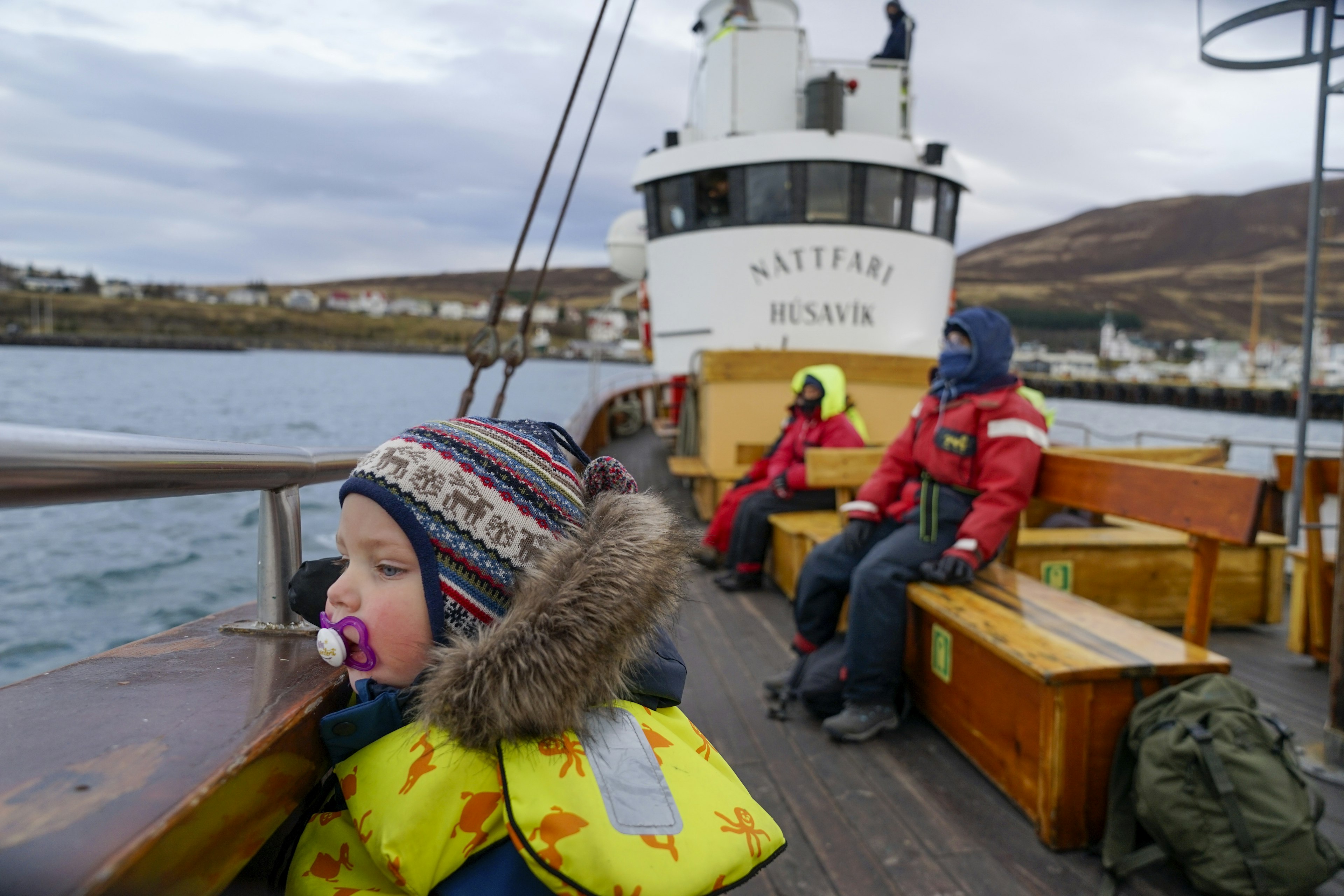 Close up of a toddler looking out in the ocean from a boat. Two people sit in the background.