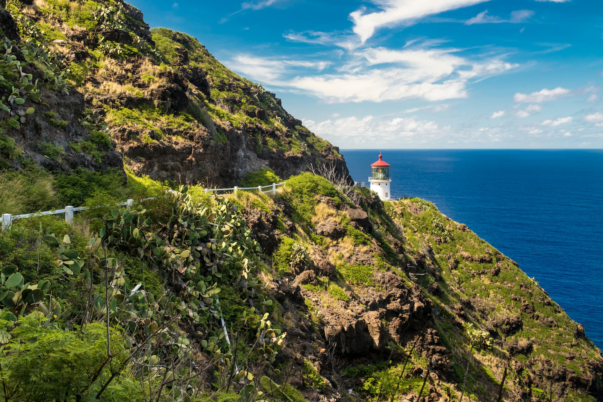 A cliff path, hugging a steep hillside, leads to a lighthouse backed by blue ocean