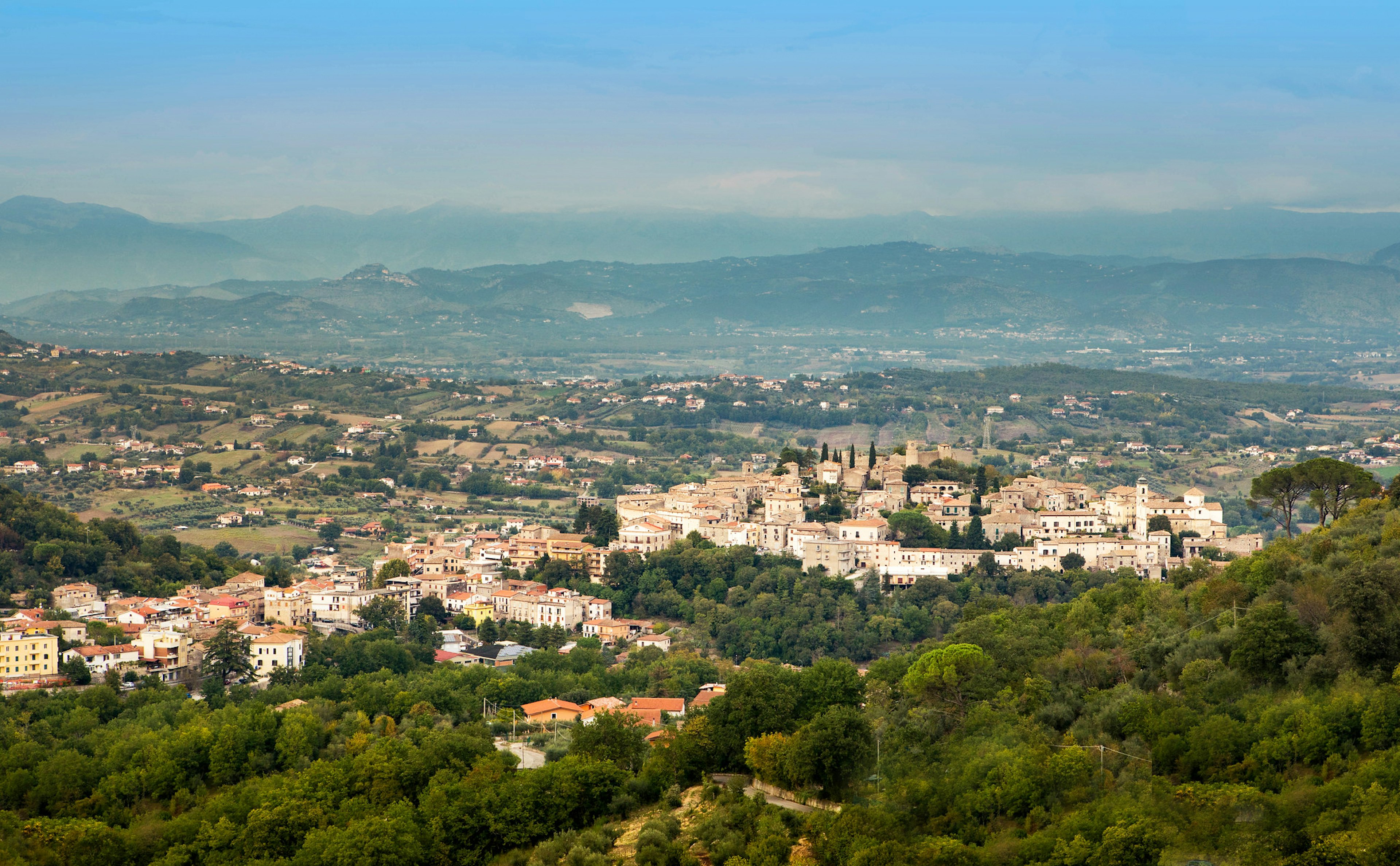 An aerial view overlooking the village of Pico in Italy shows a valley of rolling hills rimmed by blue mountains almost obscured by haze. The valley floor is dotted with cream-colored clay houses dotting fields edged in dark green trees. In the right midground is the village itself on a hill surrounded by thick walls with tall bell towers and even taller Cyprus trees