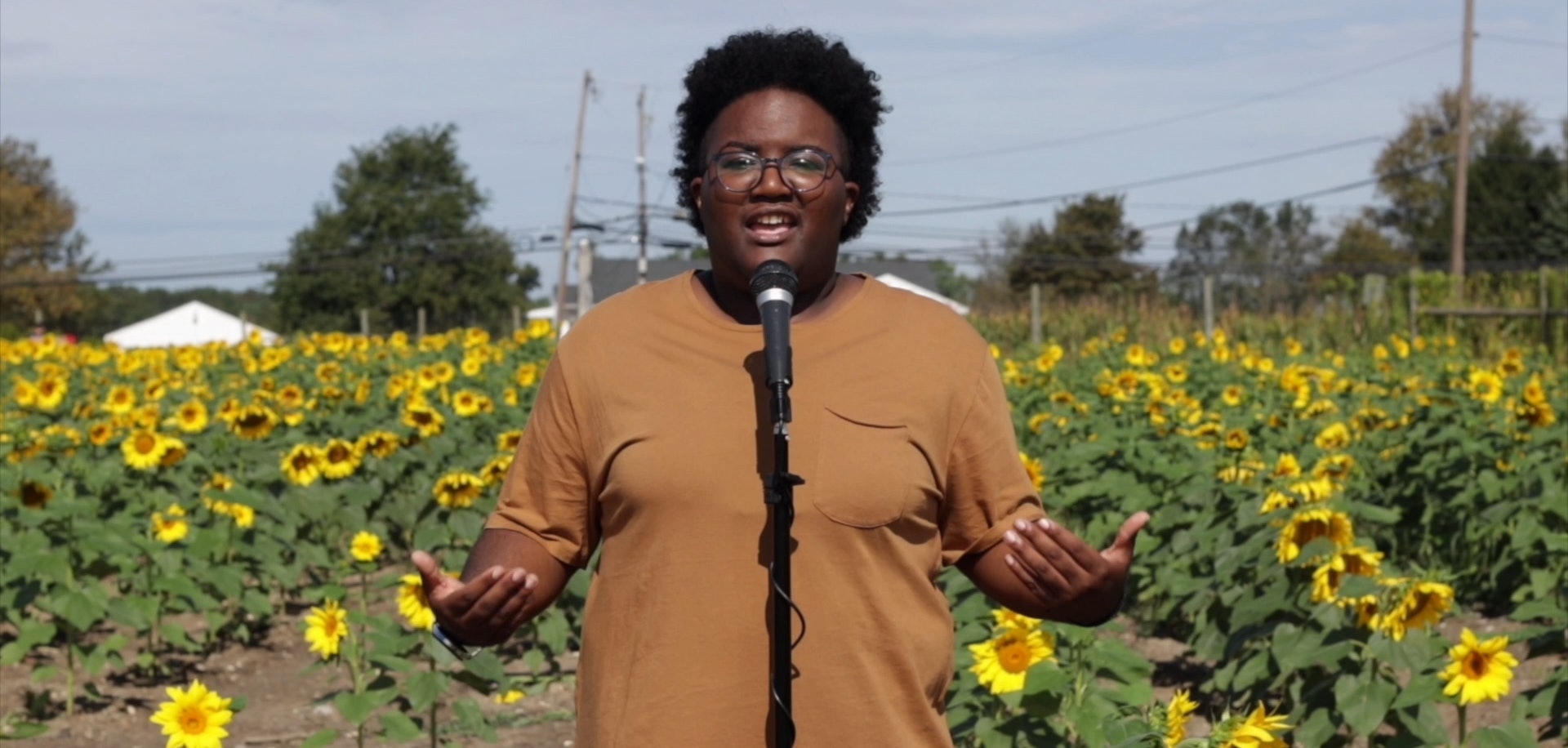 A young woman stands in front of a sunflower field on a sunny day