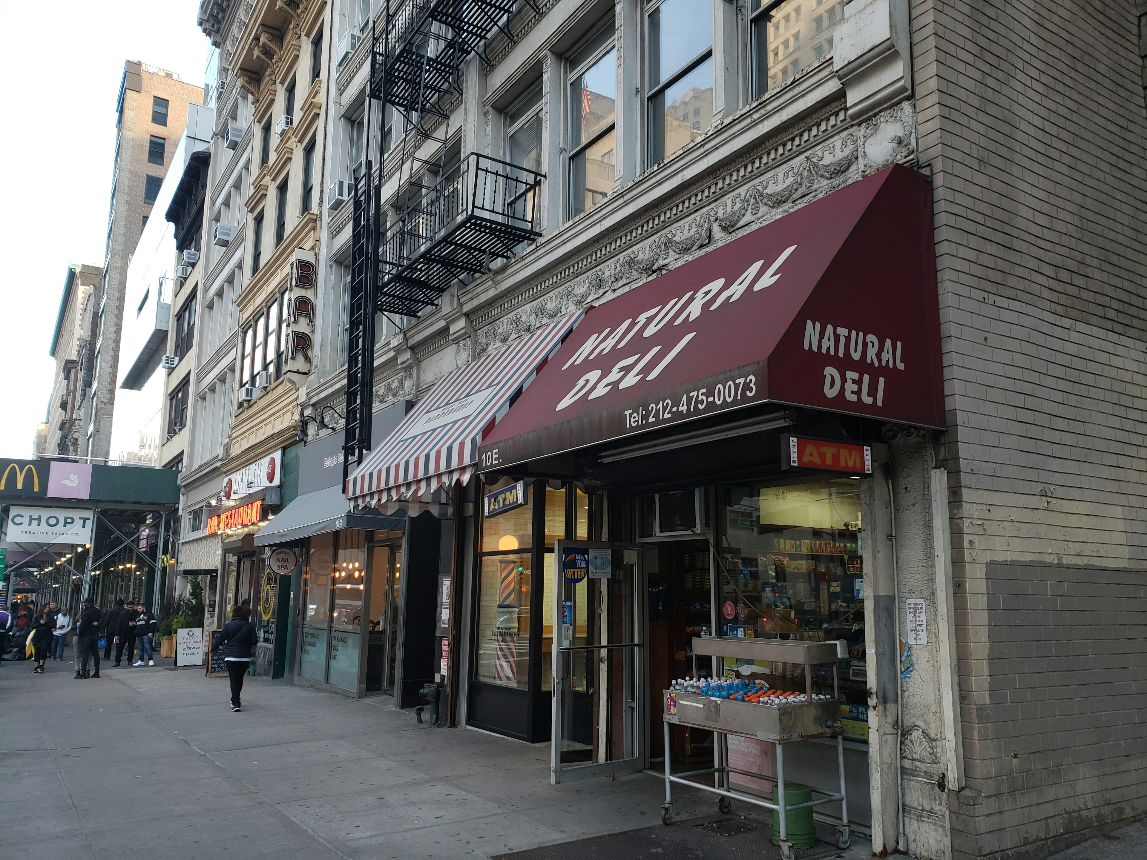 A classic New York City street lined with brick buildings and fire escapes in the Flatiron District. A maroon awning reads Natural Deli in white rounded letters in the foreground, while in the background there is a sign for a McDonald's