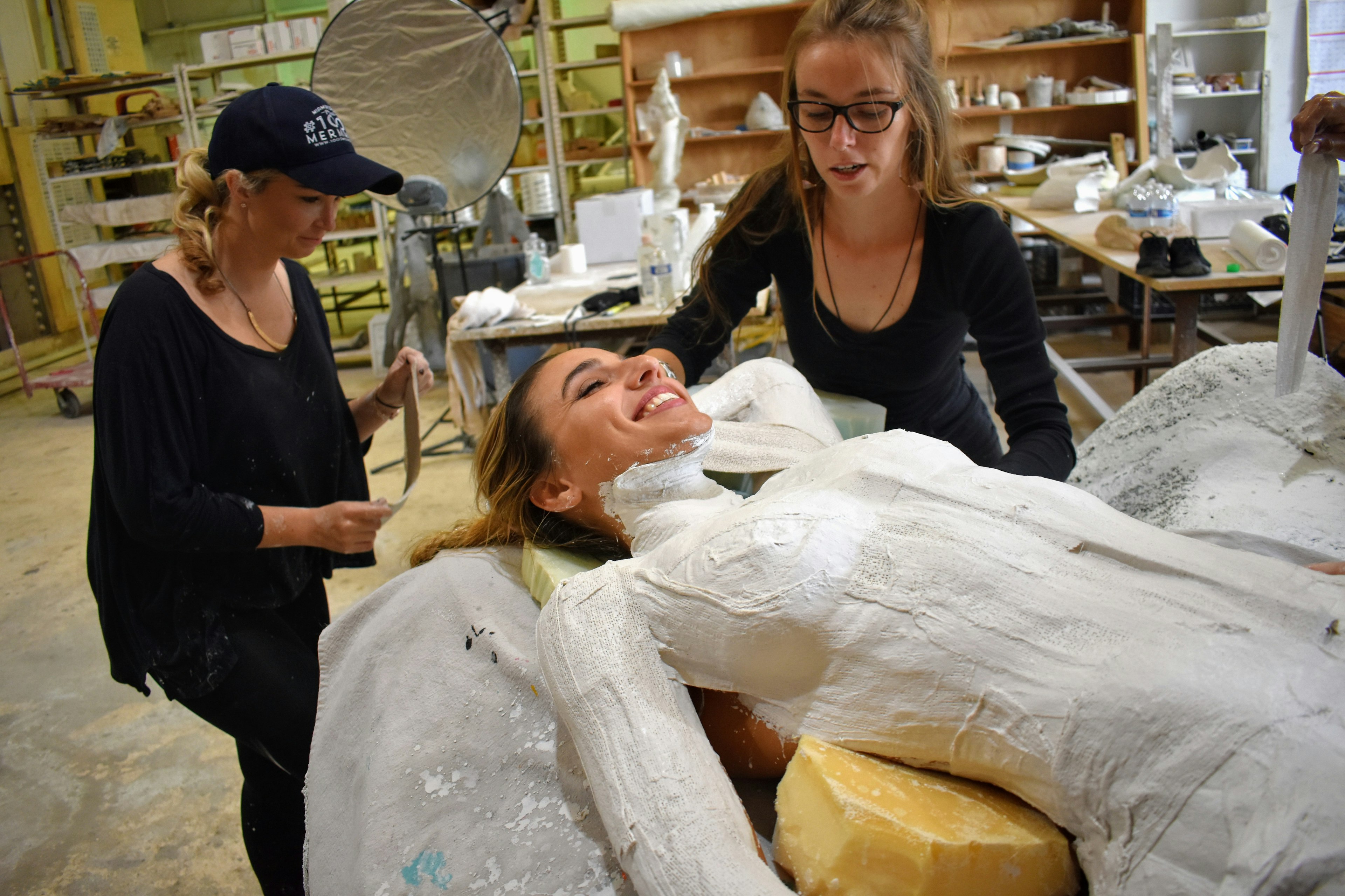 A woman covered in plaster smiles as another pair of woman place more plaster sections on her body. In the background is a collection of shelves and artist equipment scattered about on tables.