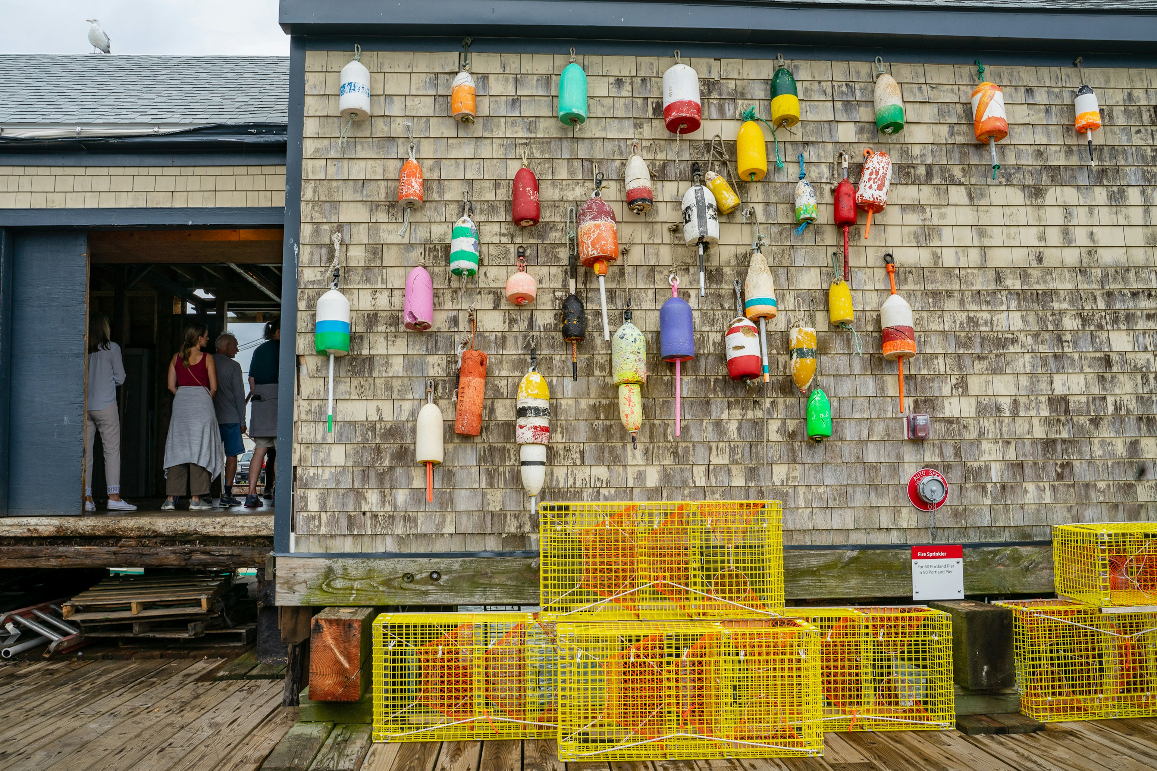 The worn wood shingles on the side of Luke's Lobster on the Portland pier is covered in colorful buoys over the wharf itself, where yellow lobster traps are stacked. To the left of the frame, up steps made from shipping palettes, stands a tour group with their backs to the camera