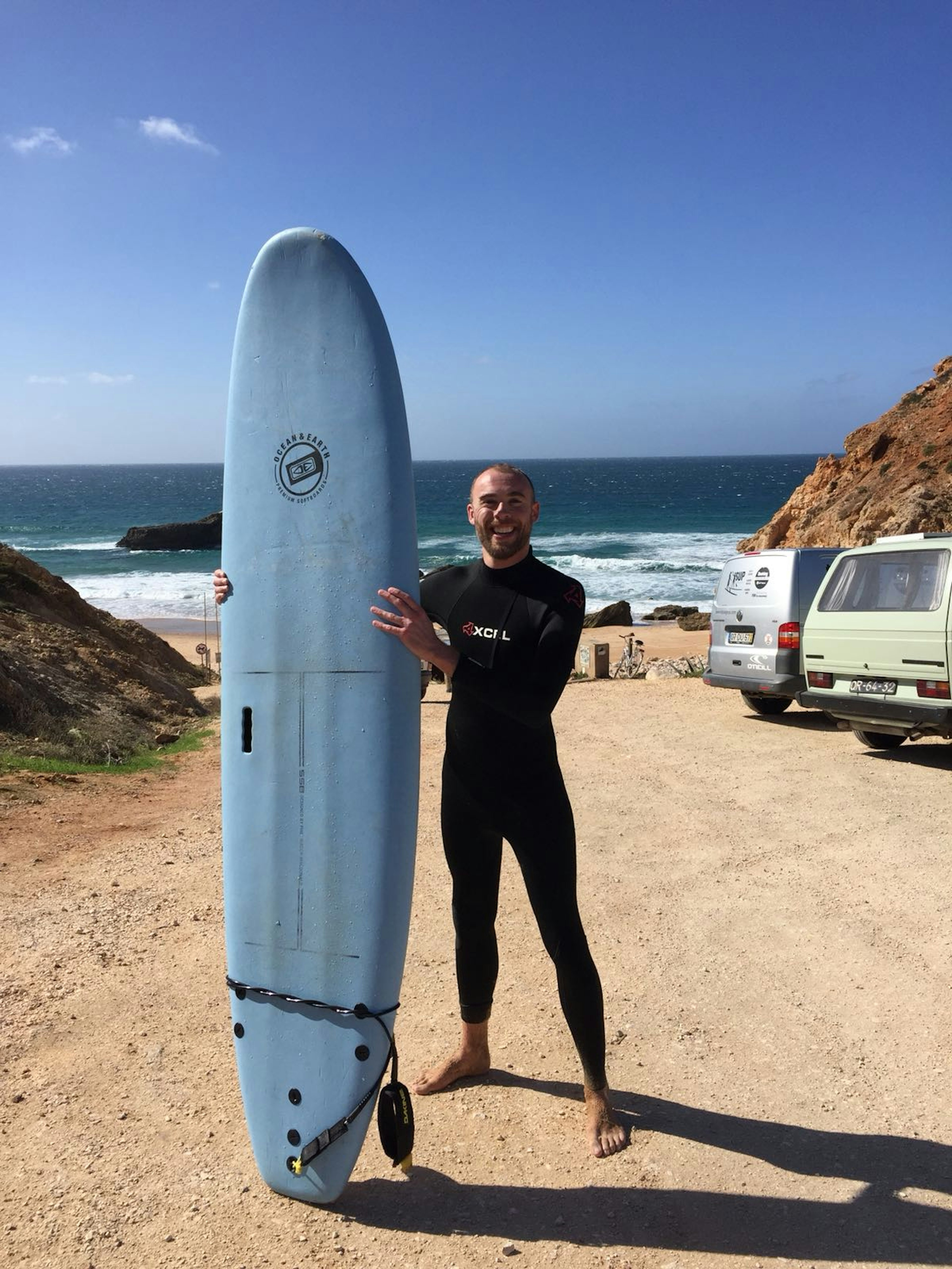 Writer Tom wears a wetsuit and holds a surfboard, with rocky cliffs, the beach and Atlantic Ocean behind him.