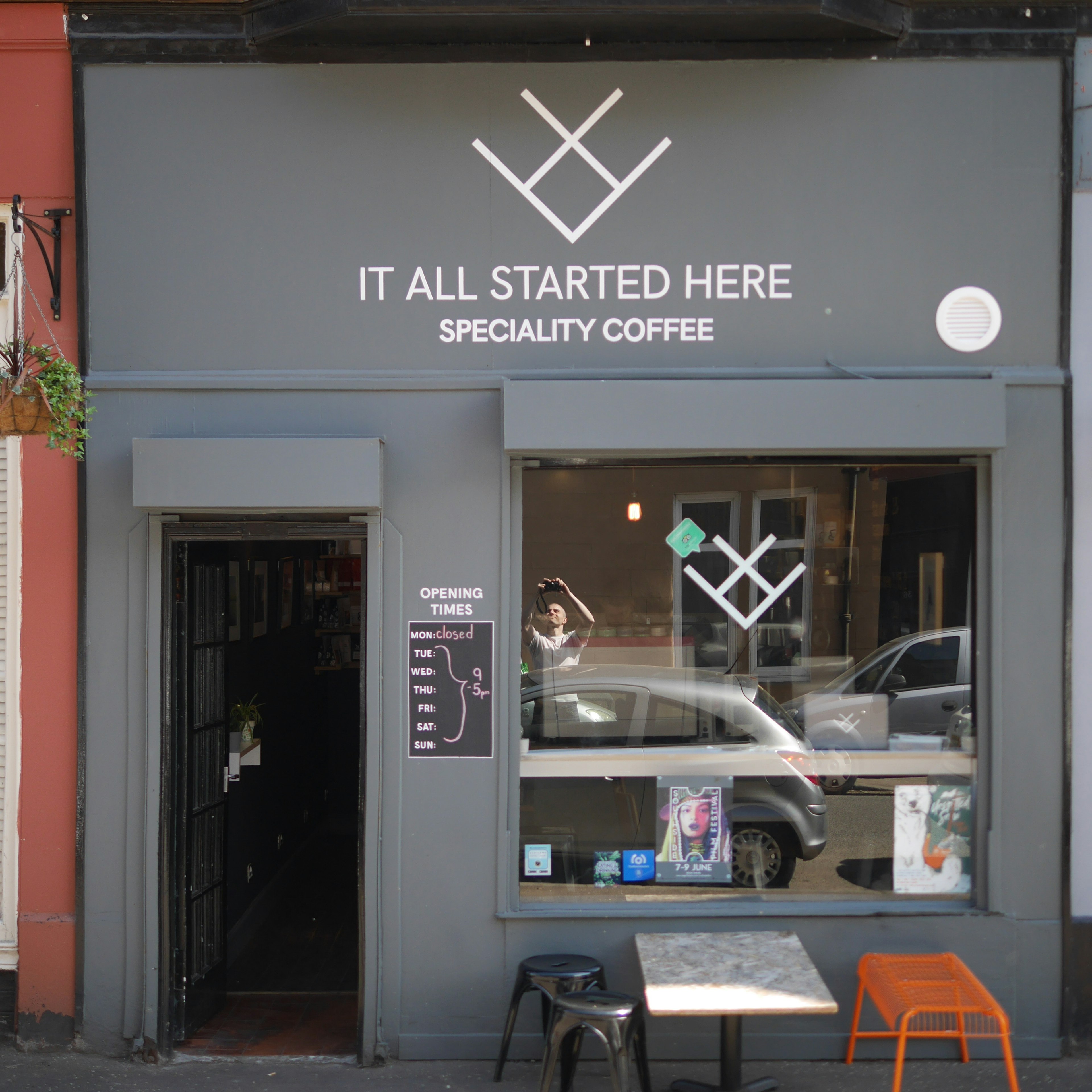 The minimalist grey facade of It All Started Here coffee shop. There is a door leading inside to the left and a large picture window to the right of it.