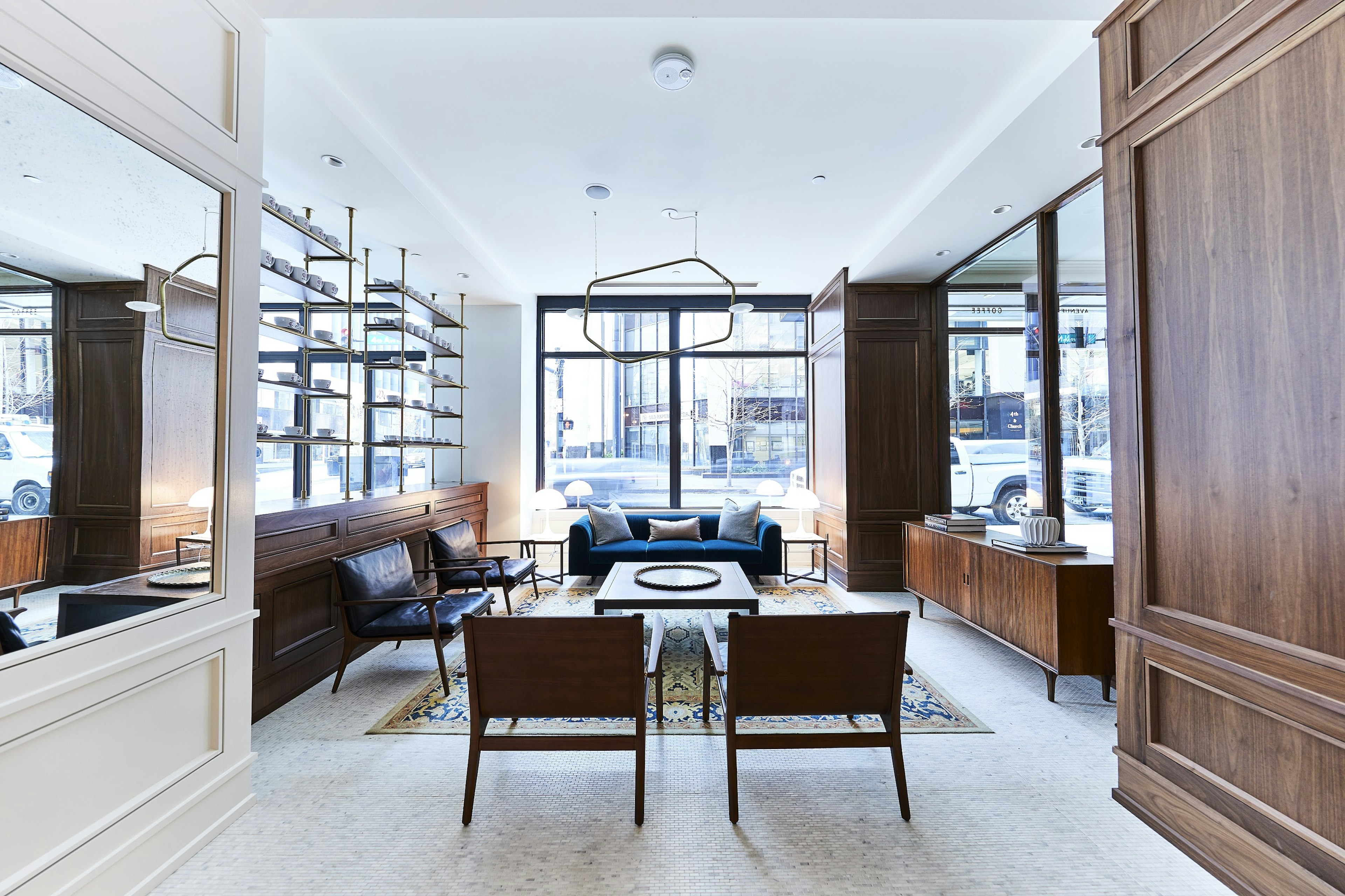 A blue velvet sofa sits at the far end of a white room full of mirrors and natural light with midcentury modern in a deep walnut tone