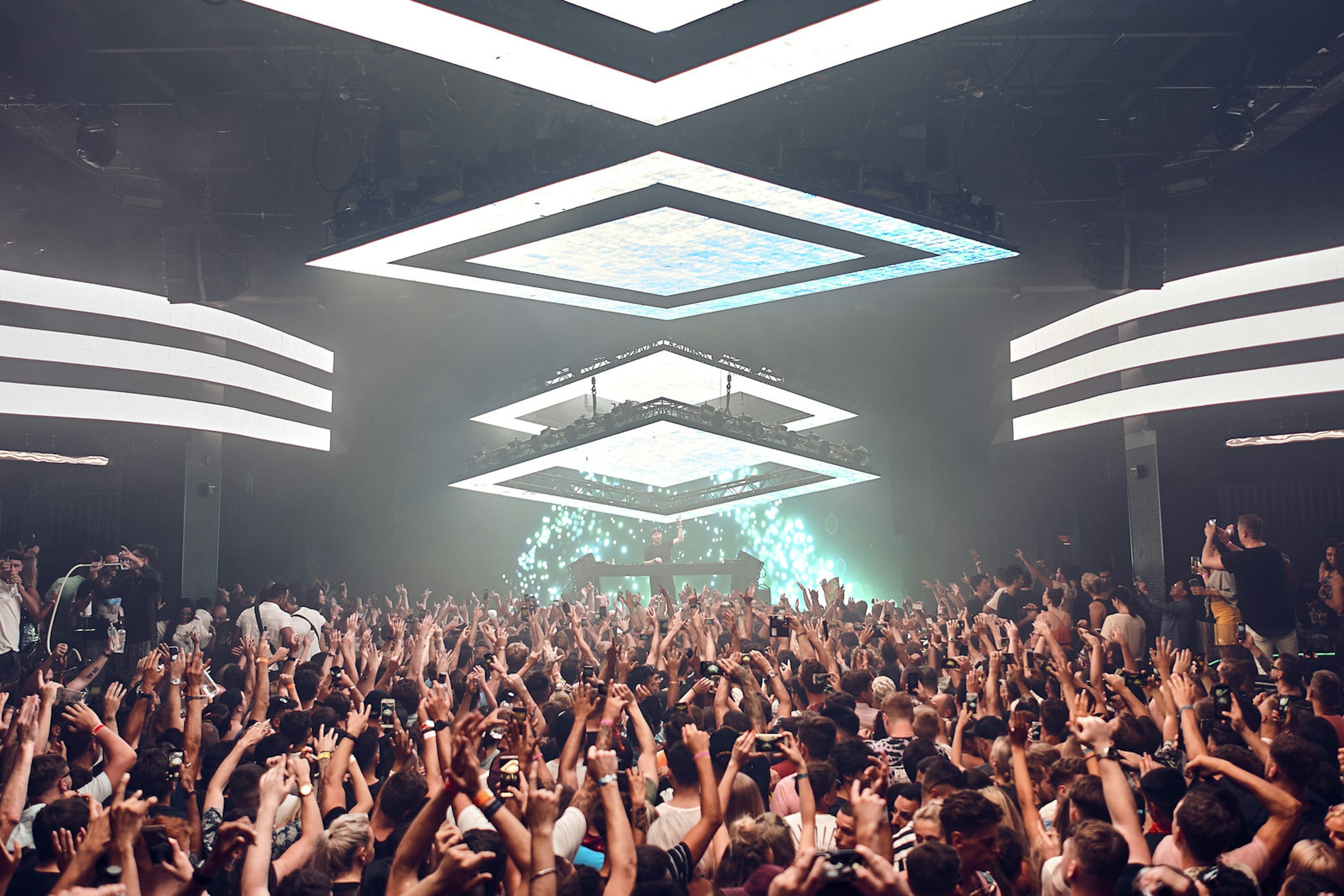 A large crowd of people dancing to the DJ Eric Prydz at megaclub Hï Ibiza. There are large white square lights on the ceiling illuminating the crowd below, who mostly have their hands in the air and backs to the camera. On stage there are green and blue lights back lighting the DJ who is wearing a black cap in front of the decks