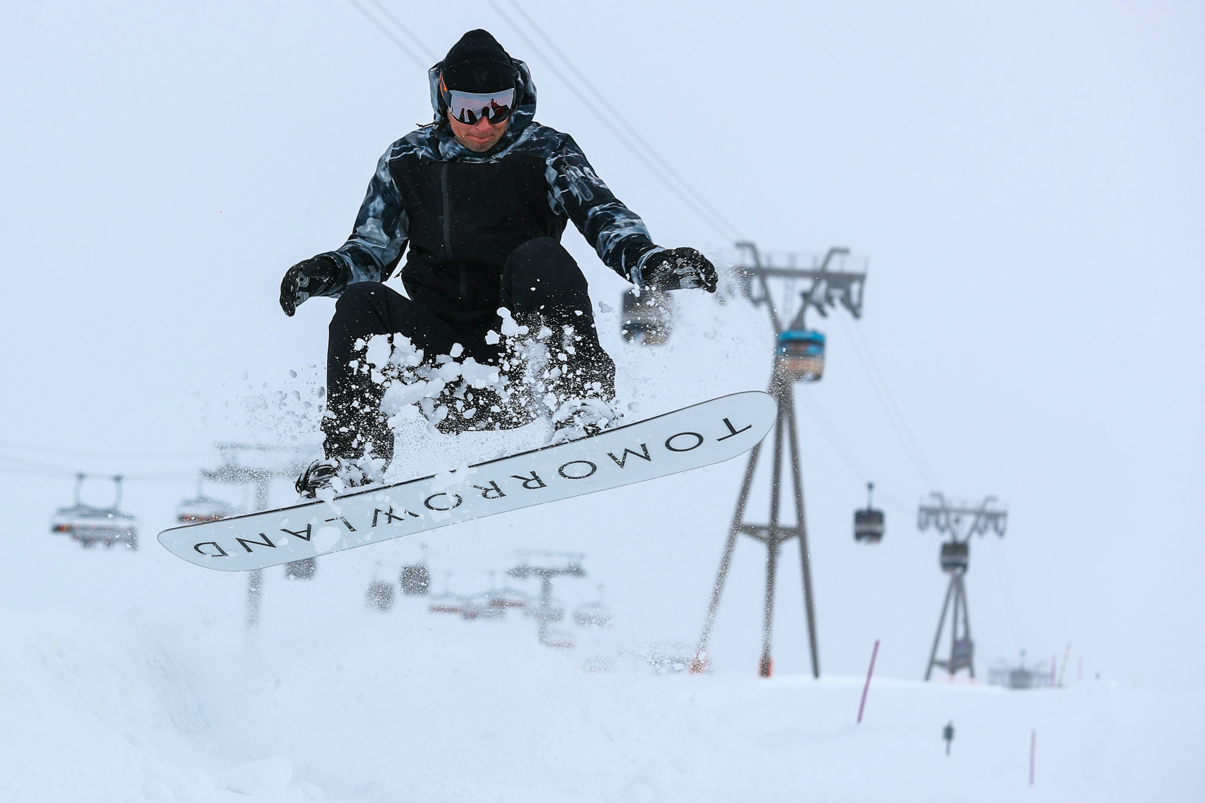 A snowboarder dressed all in black does a jump, exposing the underside of their board which reads 'TOMORROWLAND' at Tomorrowland Winter in Alpe D'Huez, France.