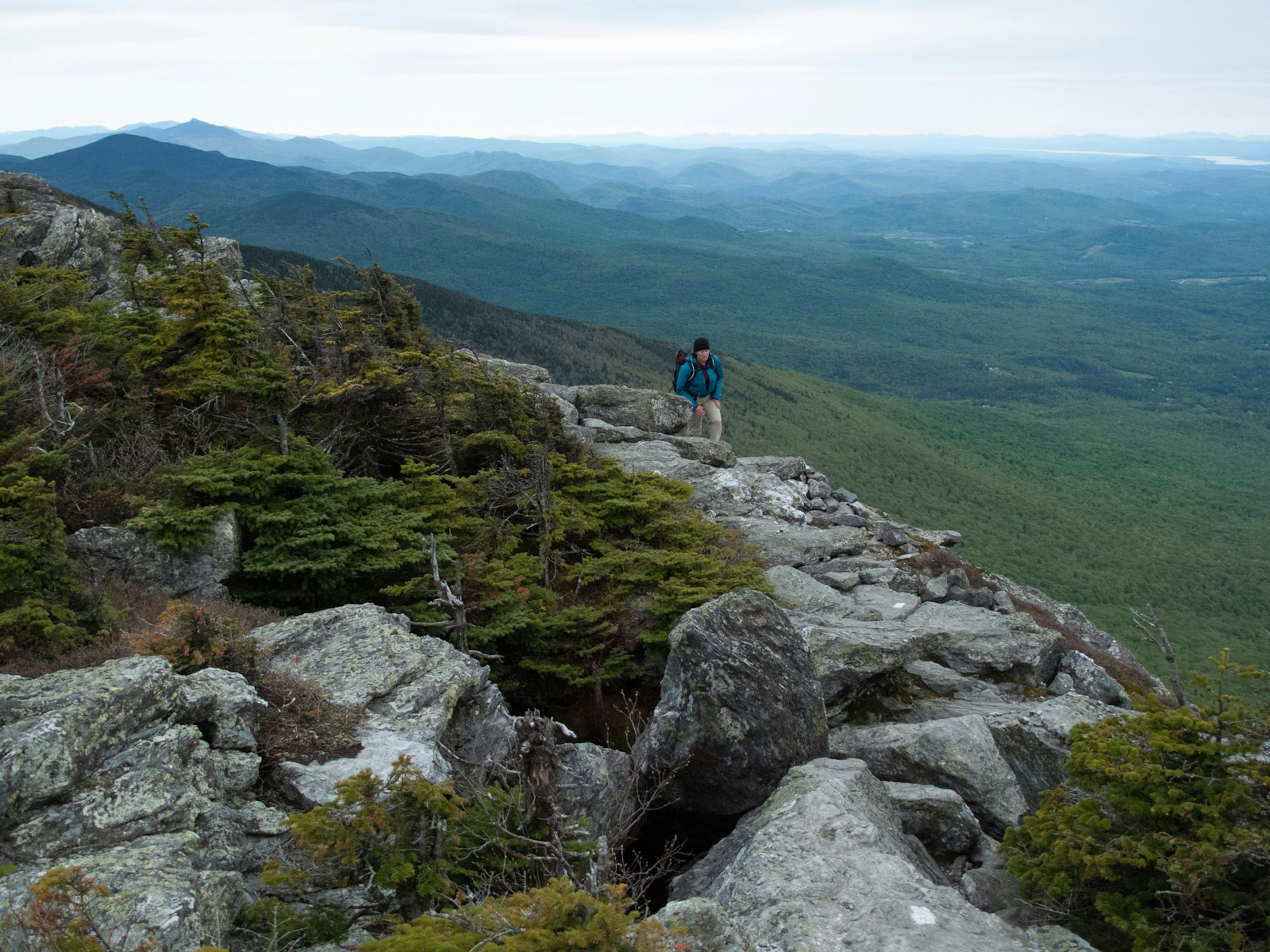 A woman walks up a rocky trail with miles of rolling green hills behind her