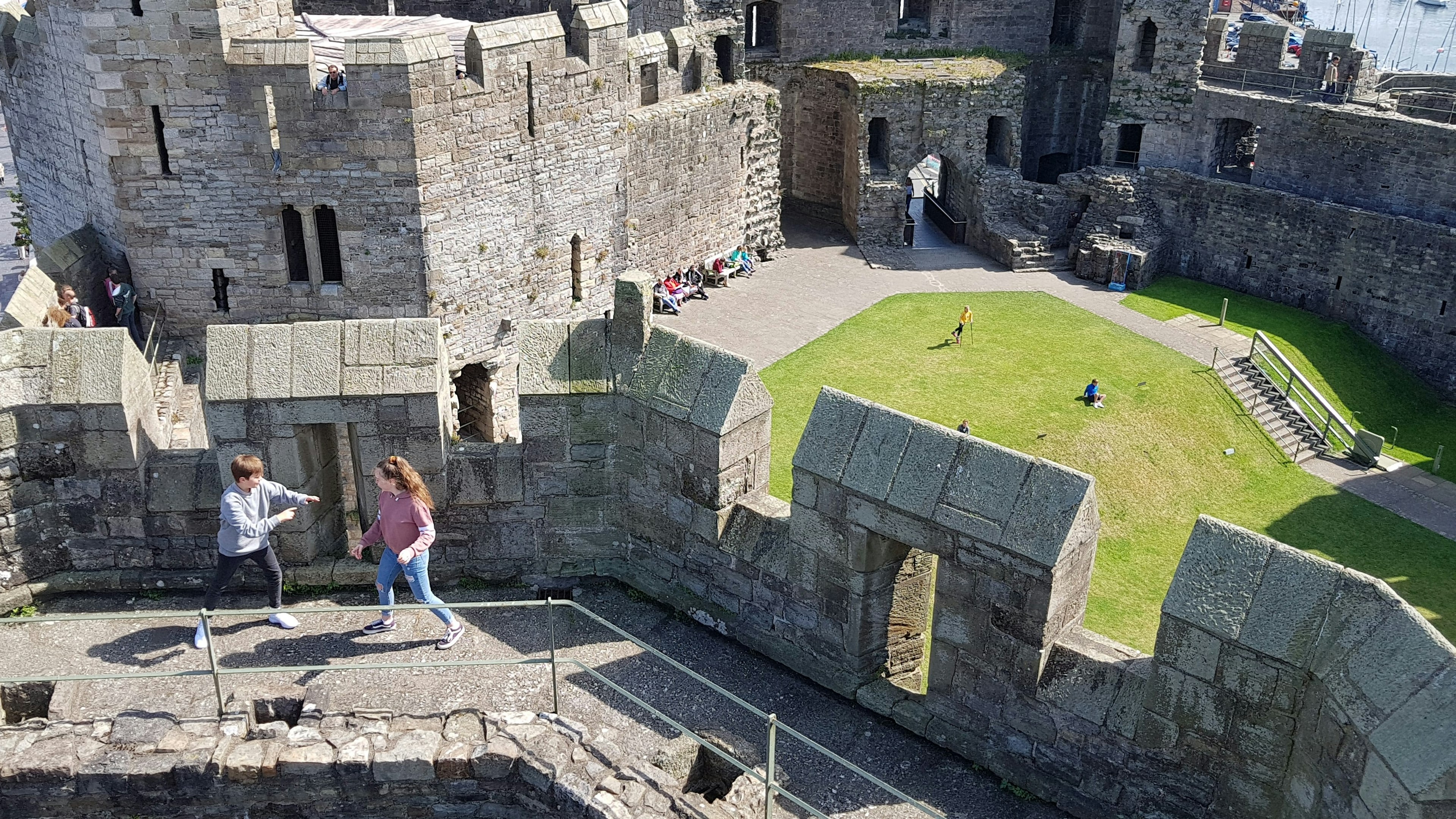 Two tweens in jeans and sweatshirts pretend to spar near stone crenelations at Caernarfon Castle in Wales. Behind them is a green lawn far below where more families play and explore. In the very far background to the right, you can just make out the masts of ships behind the castle.