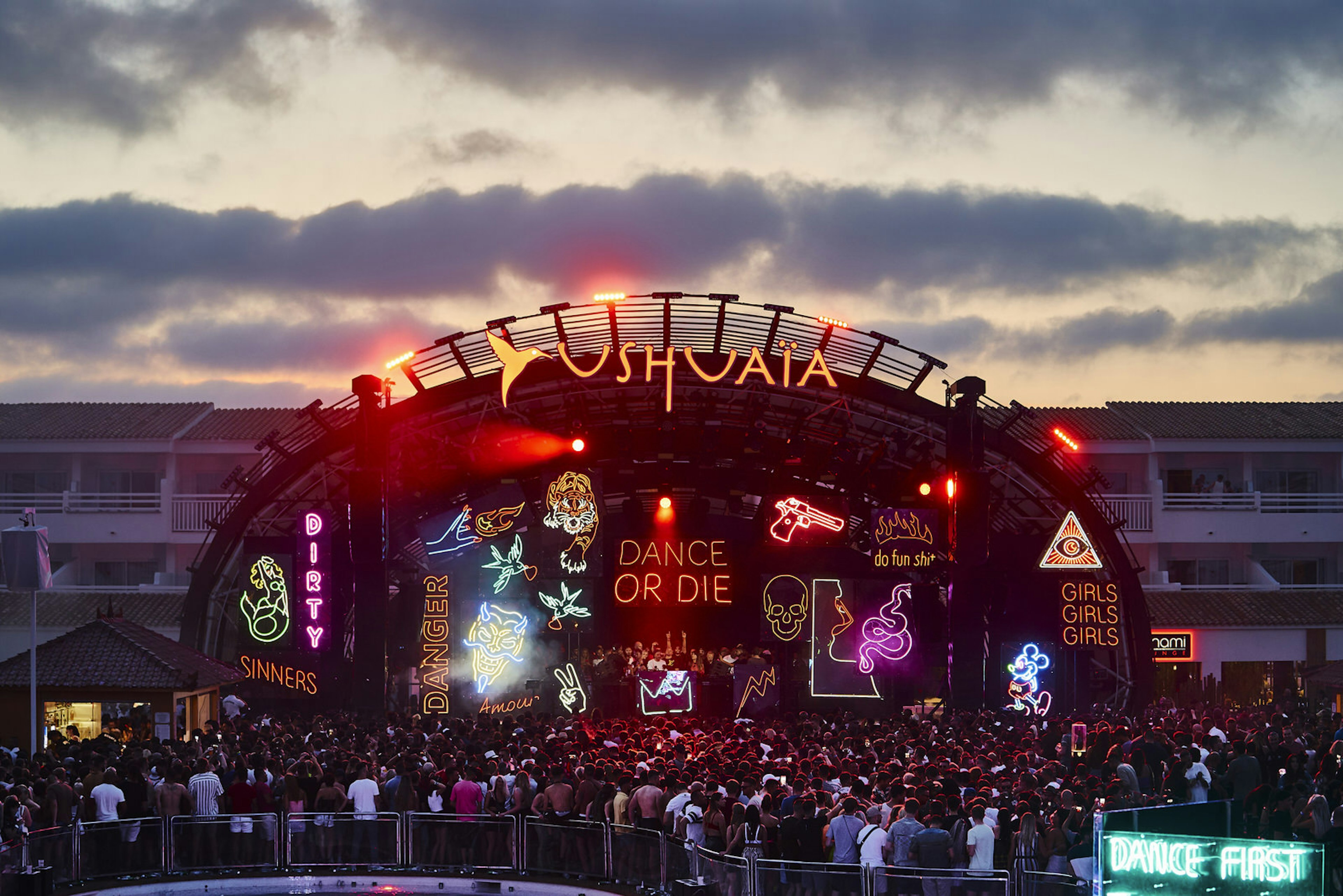 A huge crowd at Nic Fanciulli’s Dance or Die Wednesday residency at dusk. There is a huge sign with the name of the club above the outdoor stage which has several neon signs saying things like 'GIRLS GIRLS GIRLS' and 'Dance or Die'. The crowd has their backs to the camera.