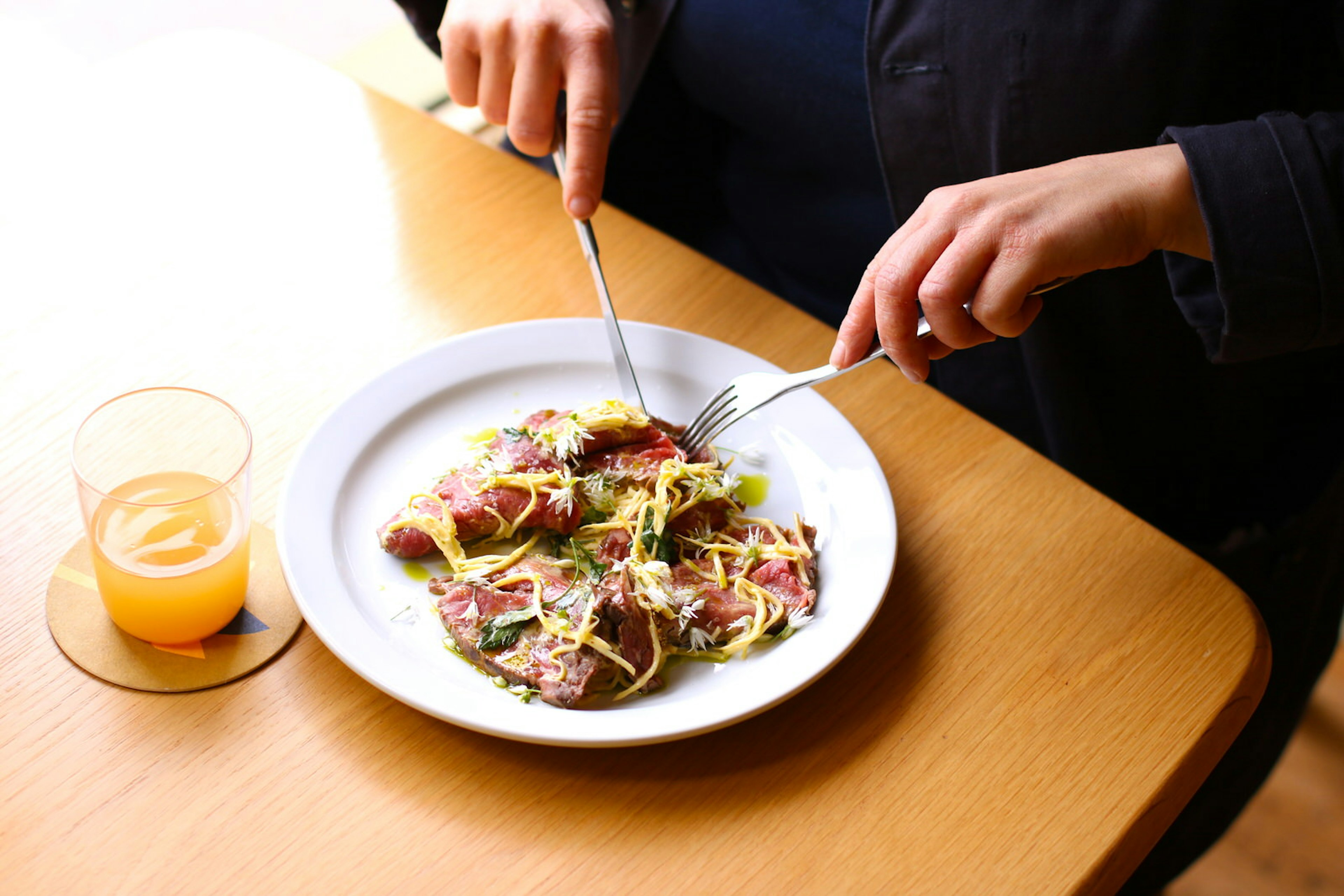 A shot of rare red meat sprinkled with oil, edible flowers and slaw. The meat is being cut by someone who is out of frame.