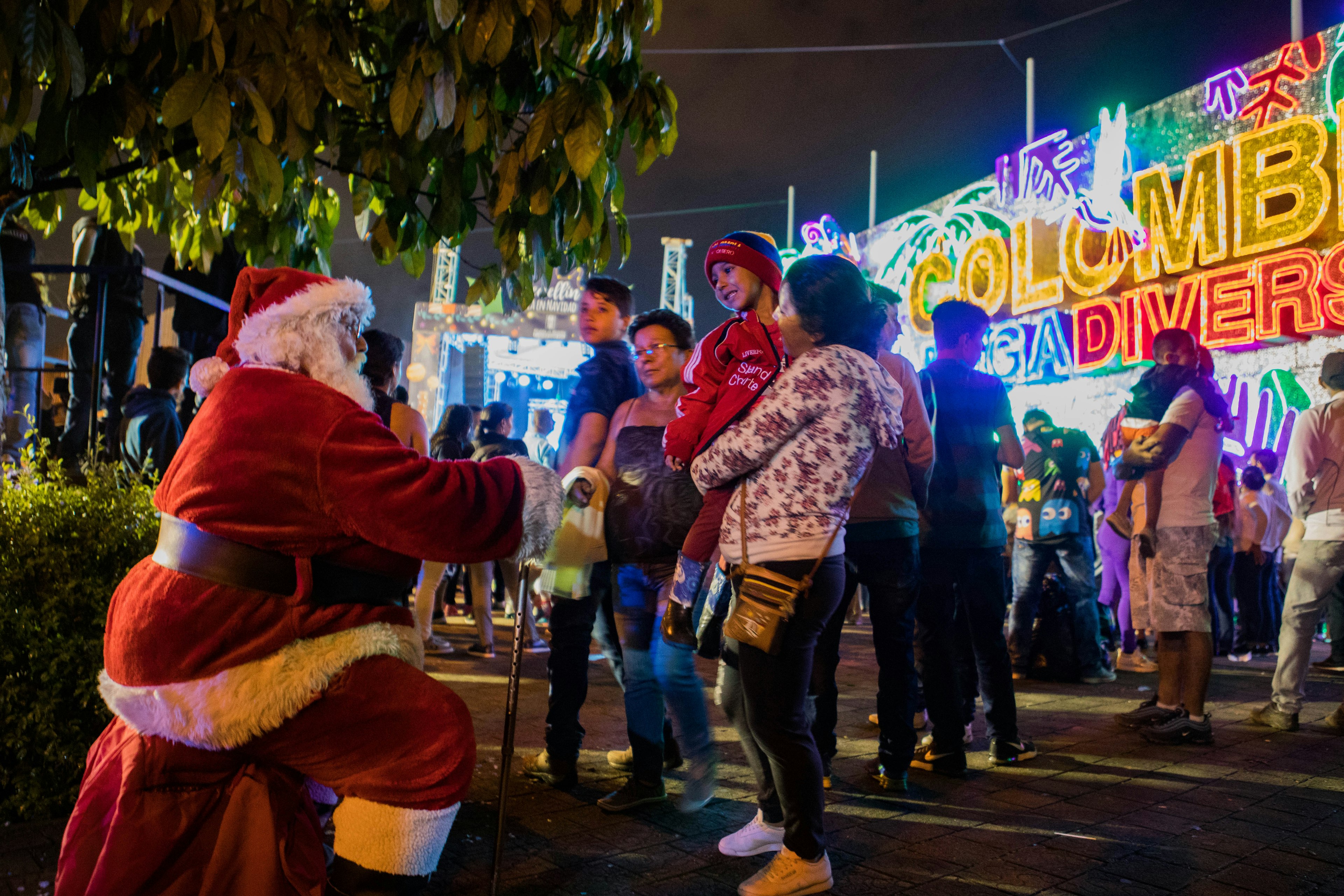 A man dressed as Santa Claus greats a child in a red, blue, and yellow striped knit cap and a red track suit held by a woman in blue jeans, a white floral sweatshirt, and other members of her family. They are in an outdoor park in front of a large brightly lit sign in yellow, blue and red