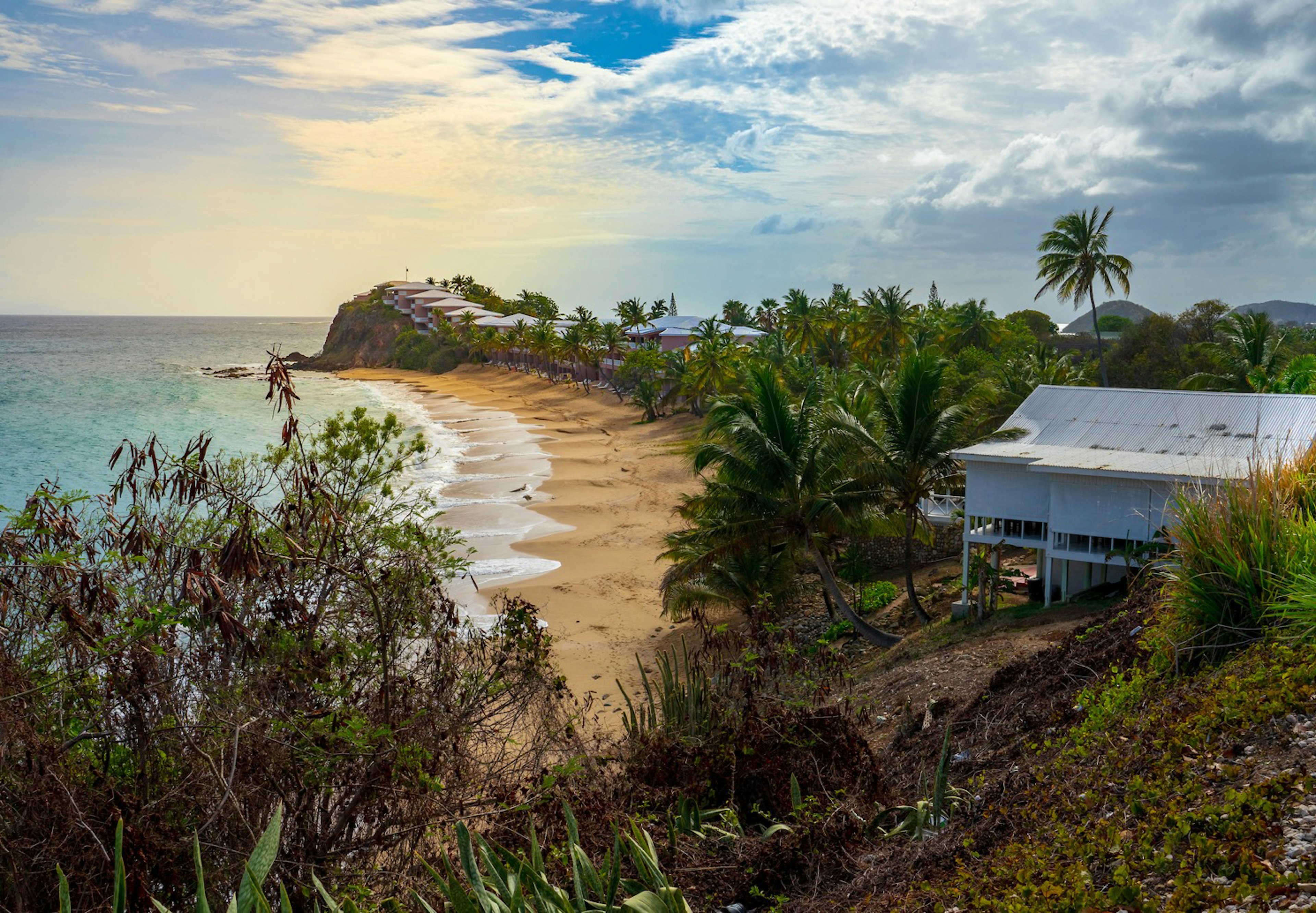 Green trees encompass an empty beach on the island of Antigua. A light blue house with a tin roof sits on stilts on a hill in Antigua and Barbuda.