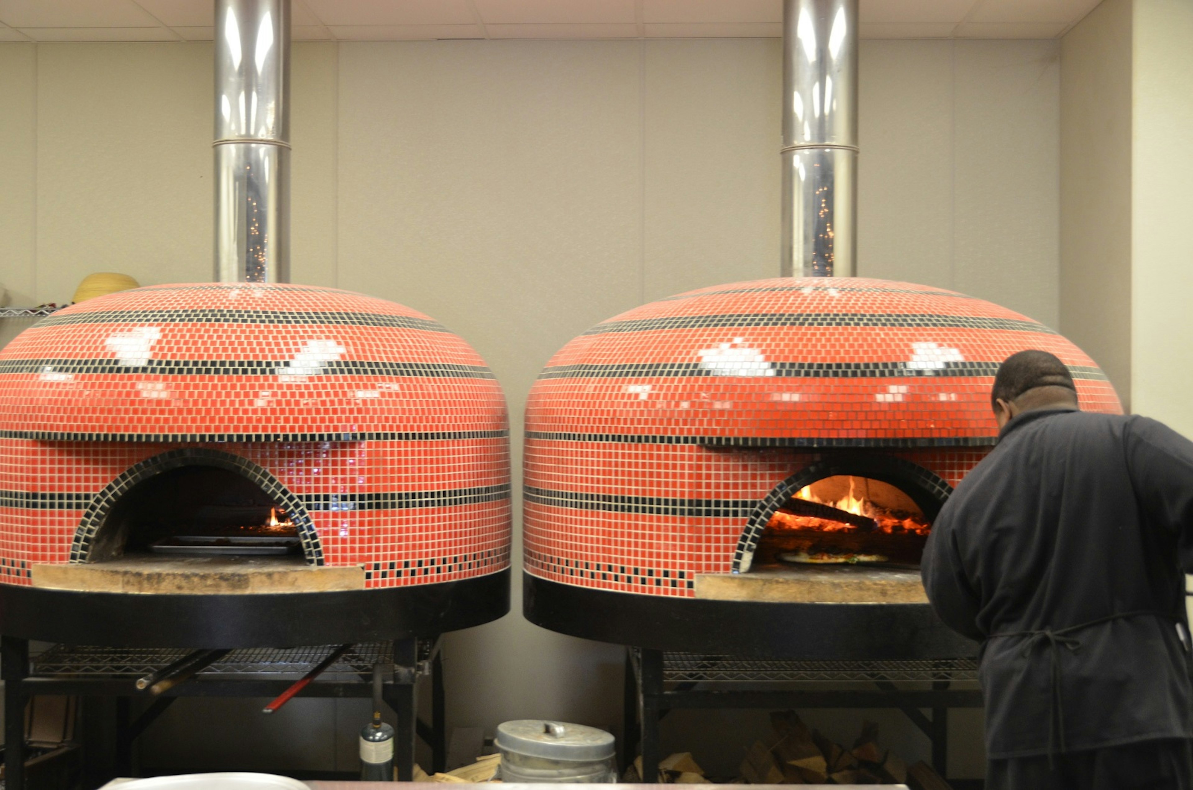 A man puts a pizza into one of two oblong ovens covered in orange and black ceramic tile.