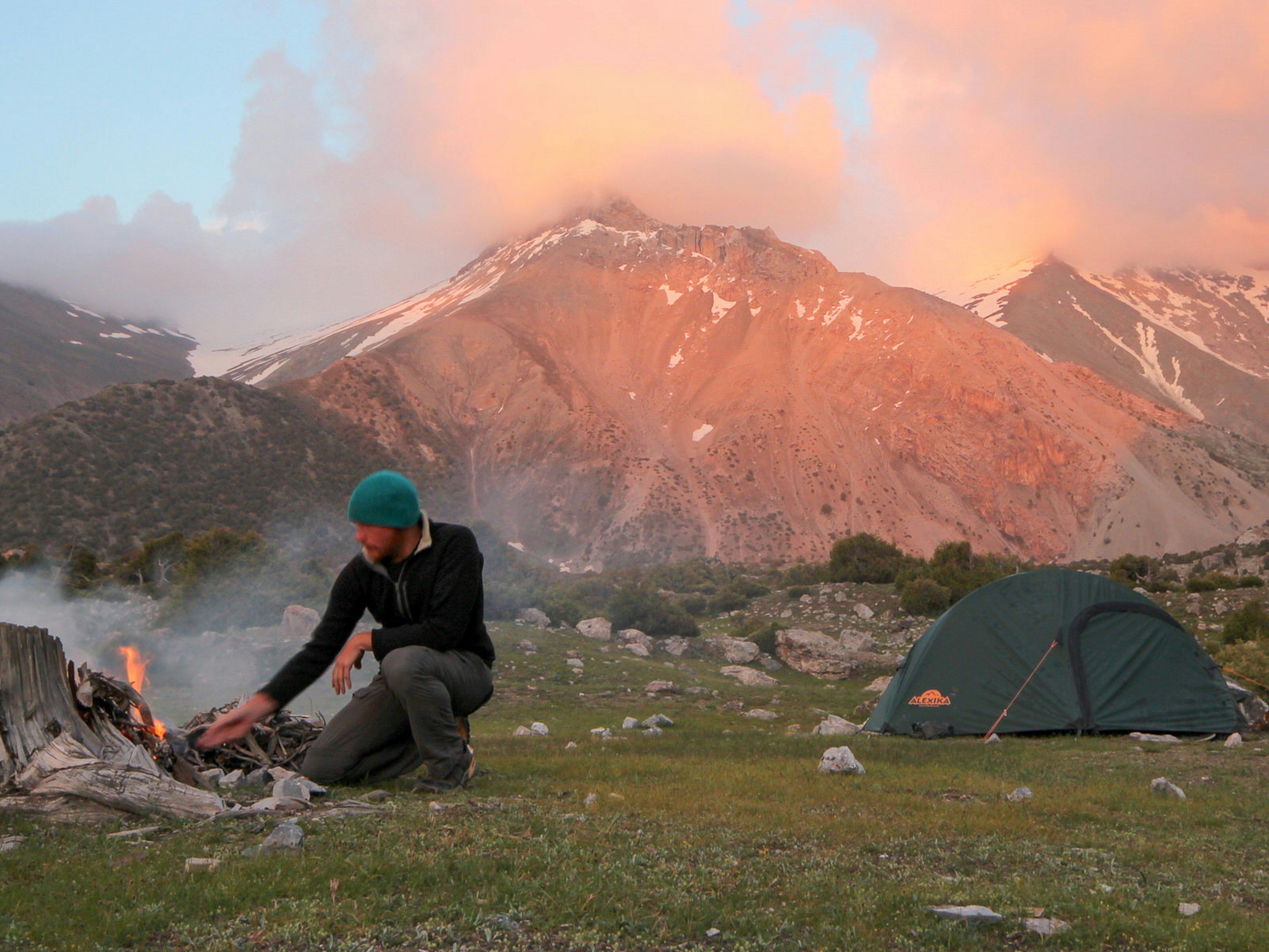 A trekker in a black shirt and green hat makes a fire from a piece of wood next to a green tent in front of mountain peaks pink from the sunset. The Fans in blushed pink whilst making camp in the Kulaikalon Basin © Stephen Lioy / Lonely Planet