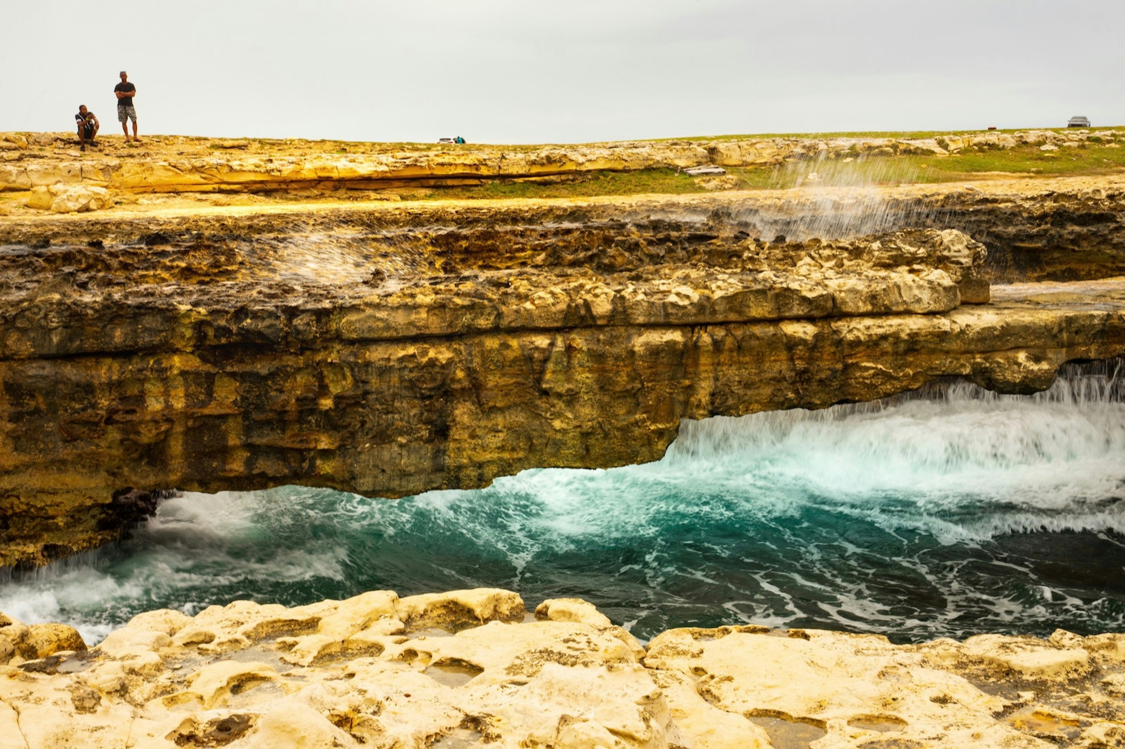 A pair of people stand on the ledge of Devil's Bridge as waves crash against the rocks in Antigua and Barbuda.