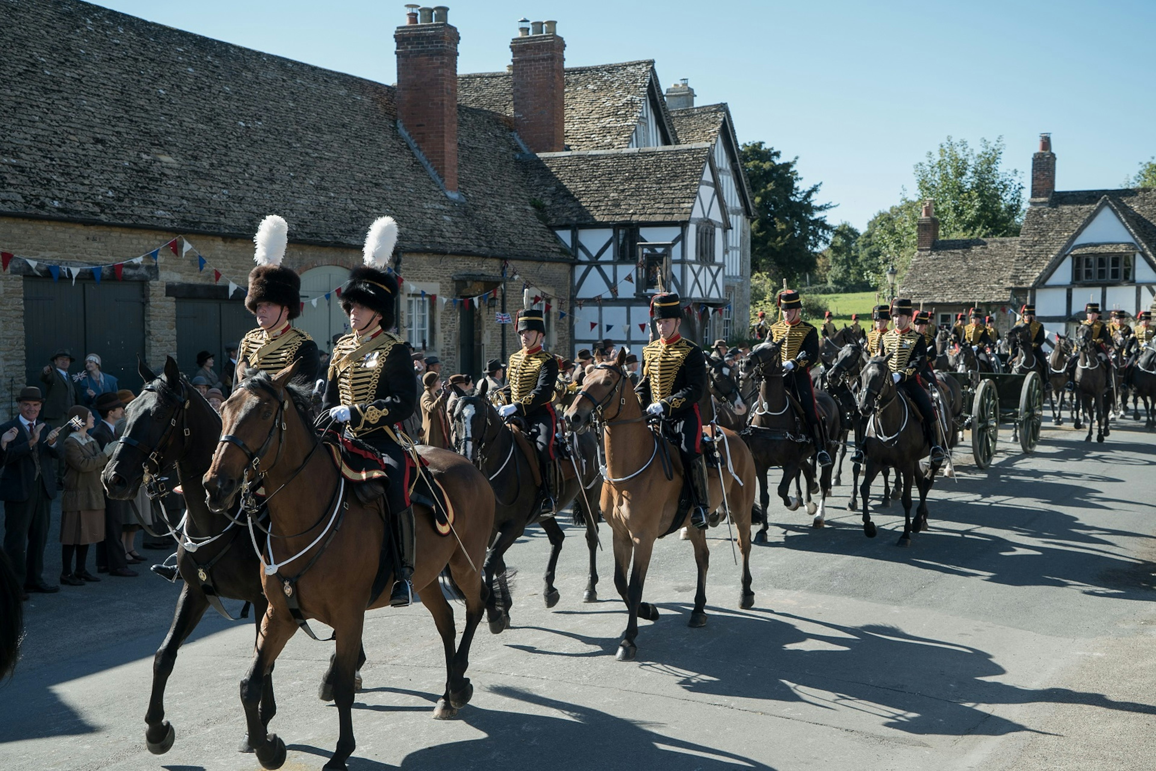 Horses and soldiers in full uniform trot through a village with people lining the street to greet them.