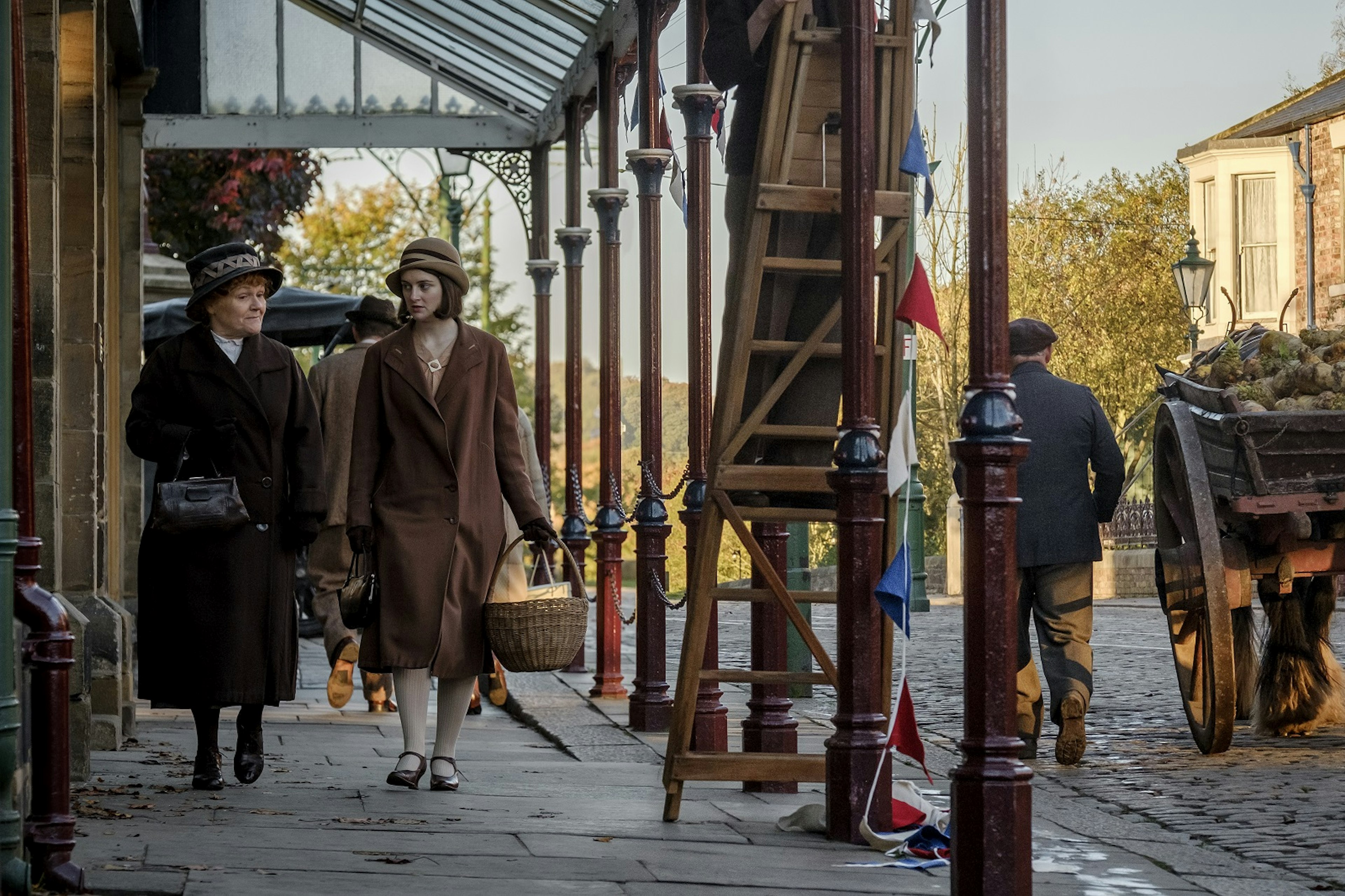 Two women (Mrs Patmore and Daisy) walk along the pavement in a village. Bunting is being put up, suggesting a party or celebration is about to take place