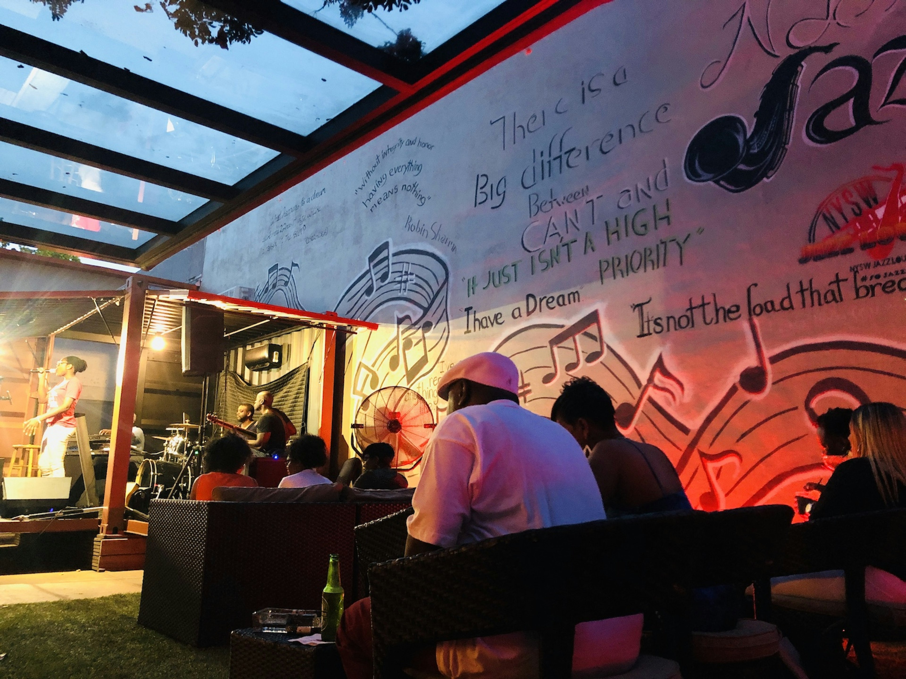 A side view of a jazz singer on the stage at NYSW Jazz Lounge. A perspex roof covers the stage and seating area next to it as patrons drink and enjoy the music. The wall next to them is covered in graffiti.