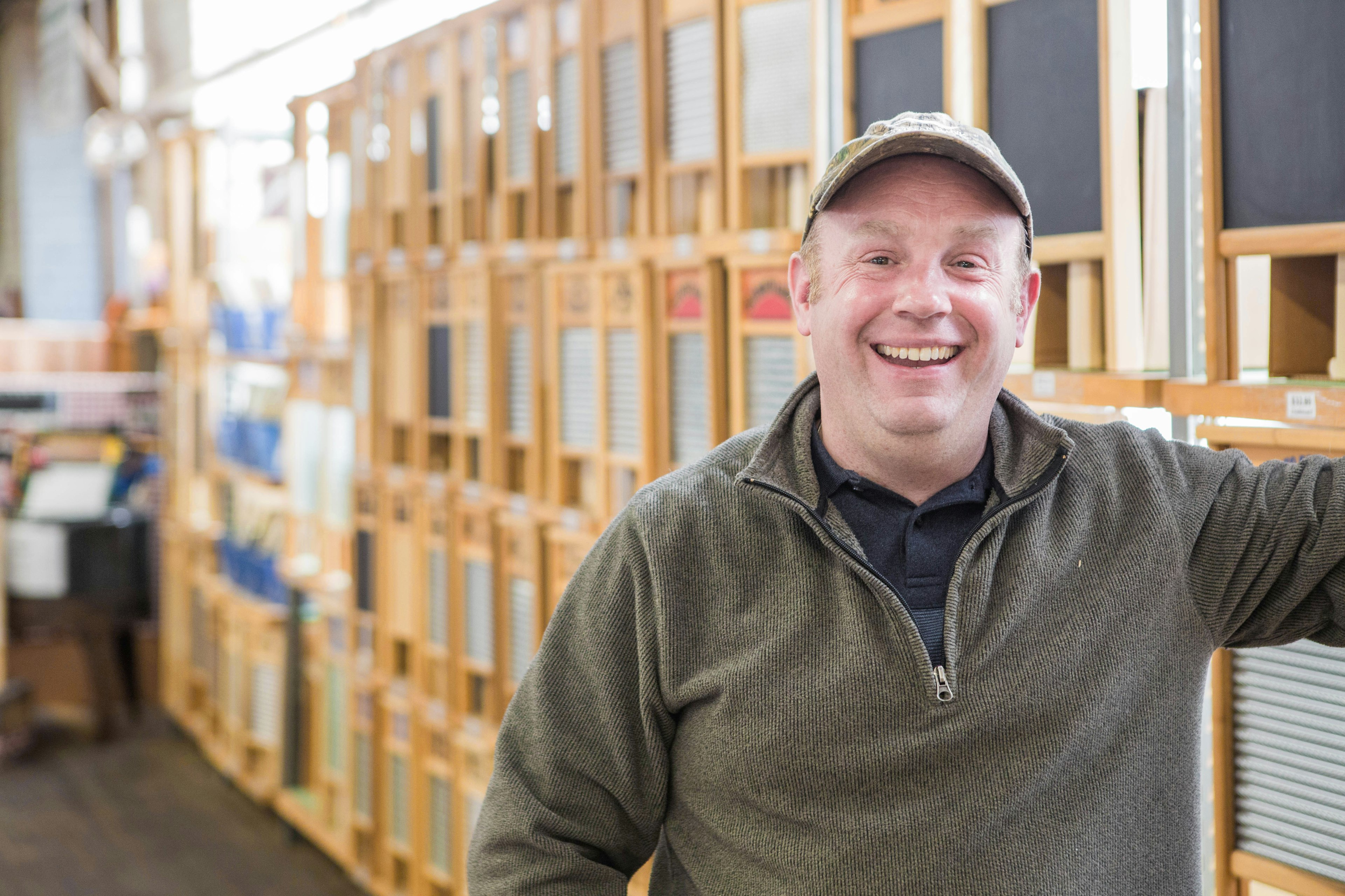 James Martin, co-owner of the Columbus Washboard Company, stands with a big cheerful smile in front of a row of washboards. He wears an olive green quarter-zip pullover with a grey polo shirt underneath and a tan ball cap.