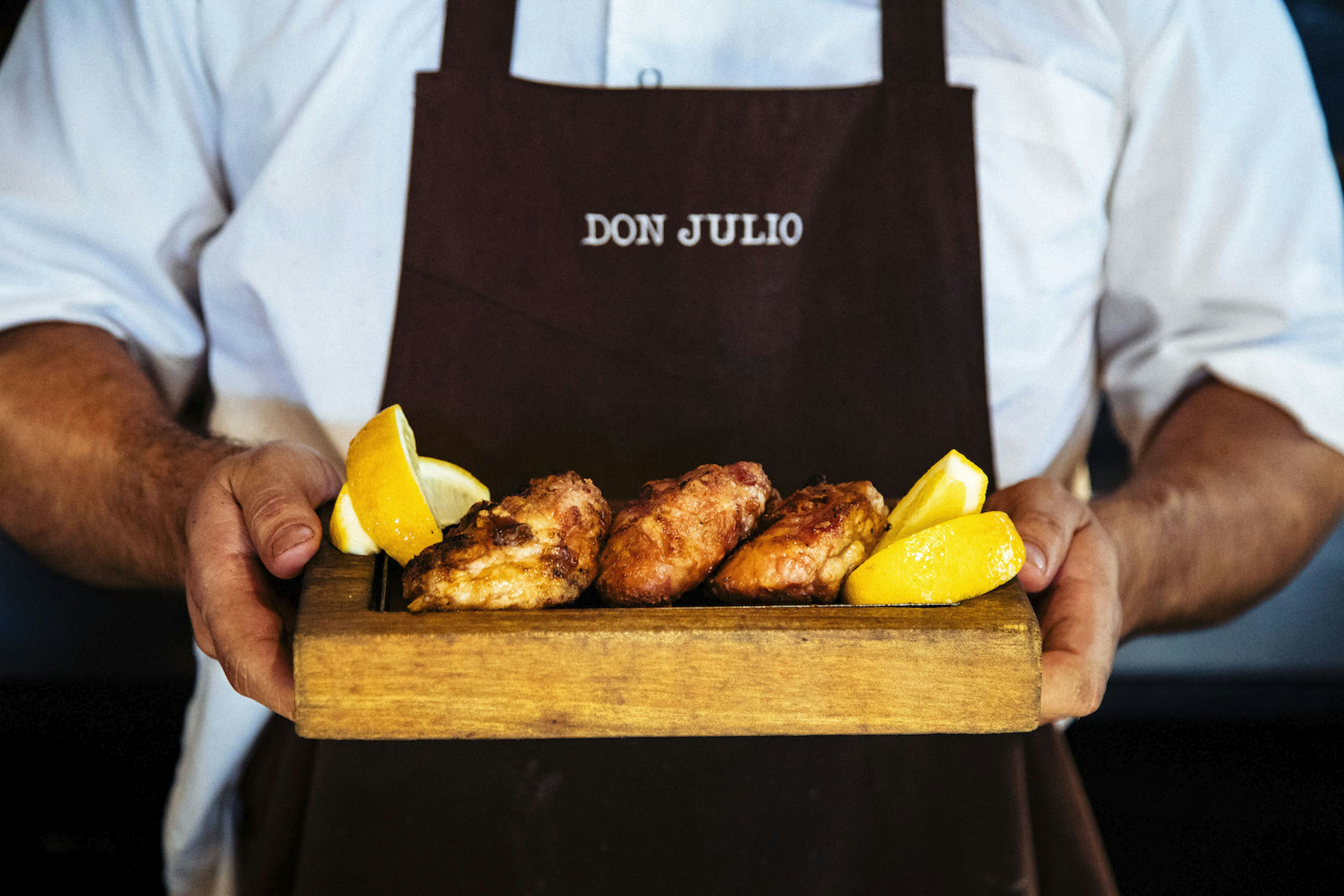 A waiter holding a fried meat dish on a rectangular wooden plate with two lemon wedges crossed on the corner