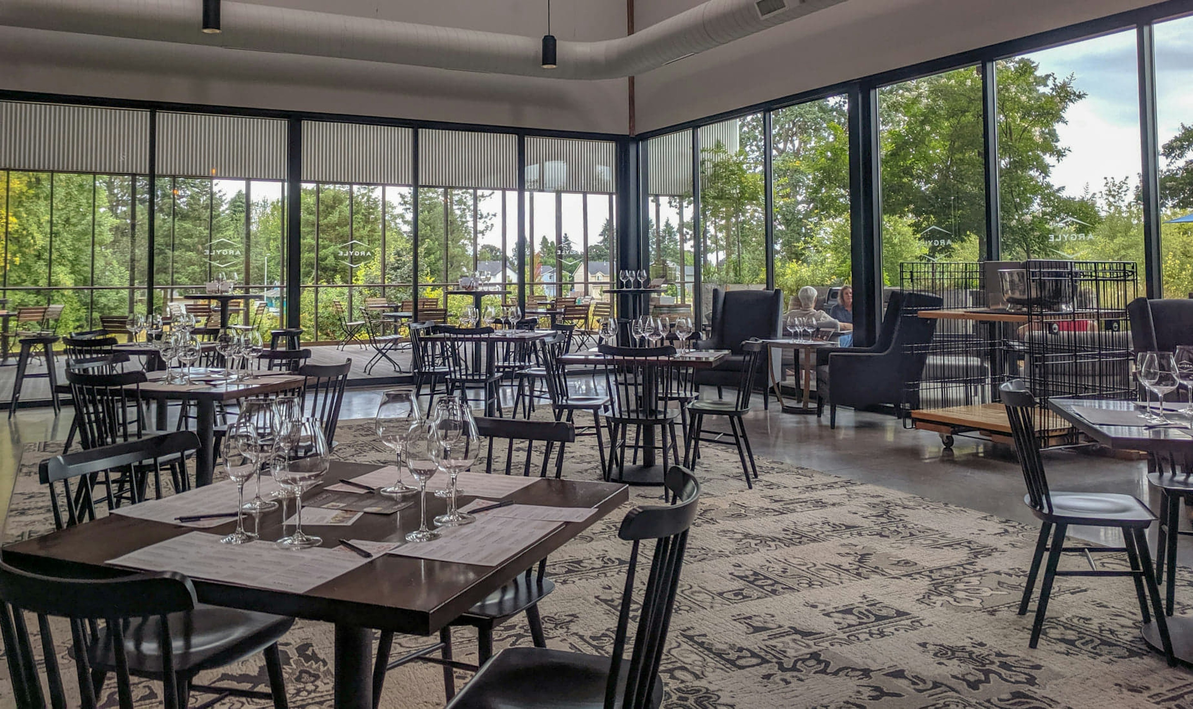 The spacious tasting room at Argyle Winery is made up of glass walls on two sides overlooking green trees and a roomy patio. Inside, black minimalist Quaker-style chairs are arranged around brown square dining tables, each set with clusters of three or four wine glasses and tasting menus. A beige and black carpet with a simple, abstract design covers the dark wood floor. Above the windows, the walls are white with simple black canister pendant lamps