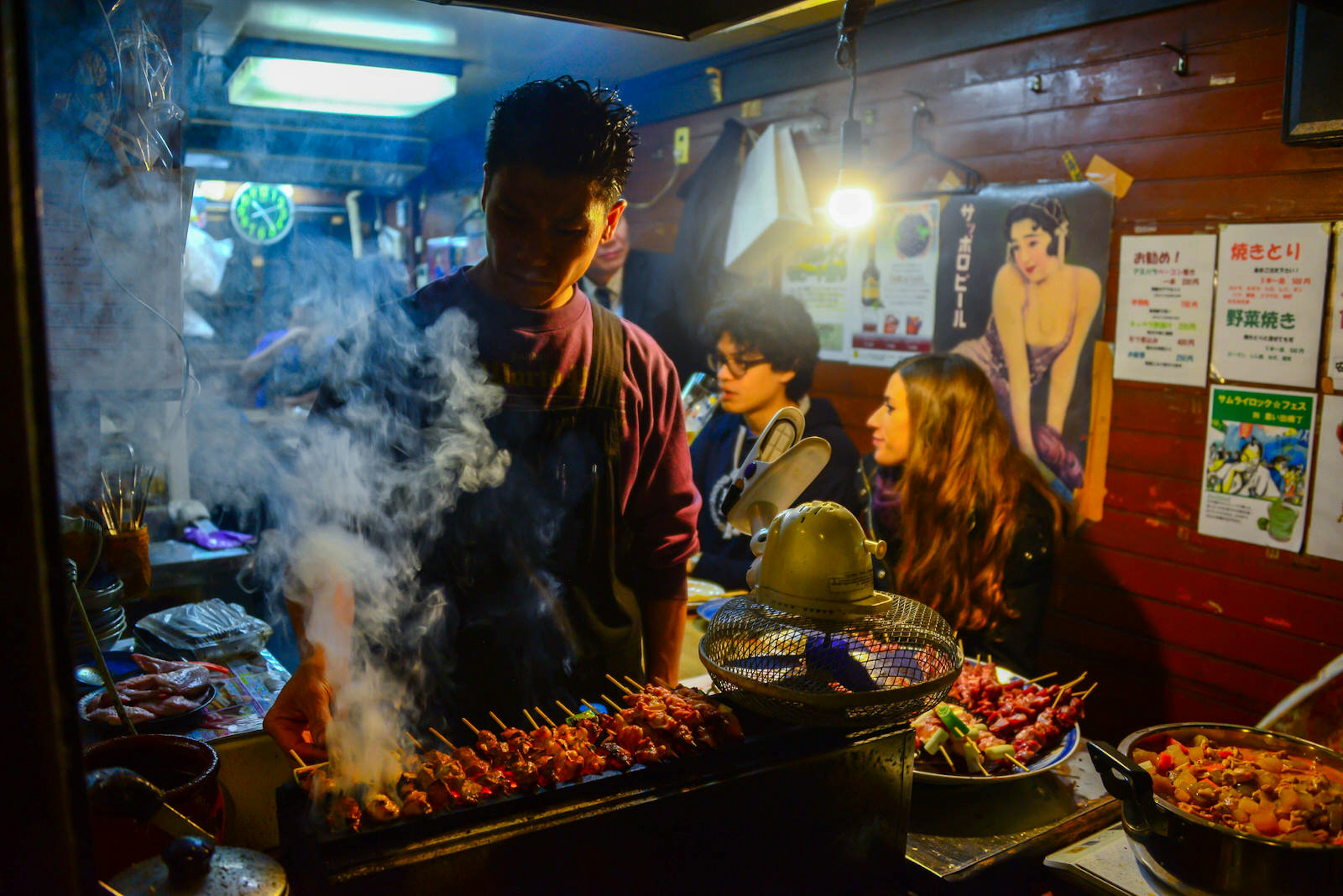 A Japanese man grills BBQ meat for customers waiting in the background at a small food stall at Omoide Yokocho