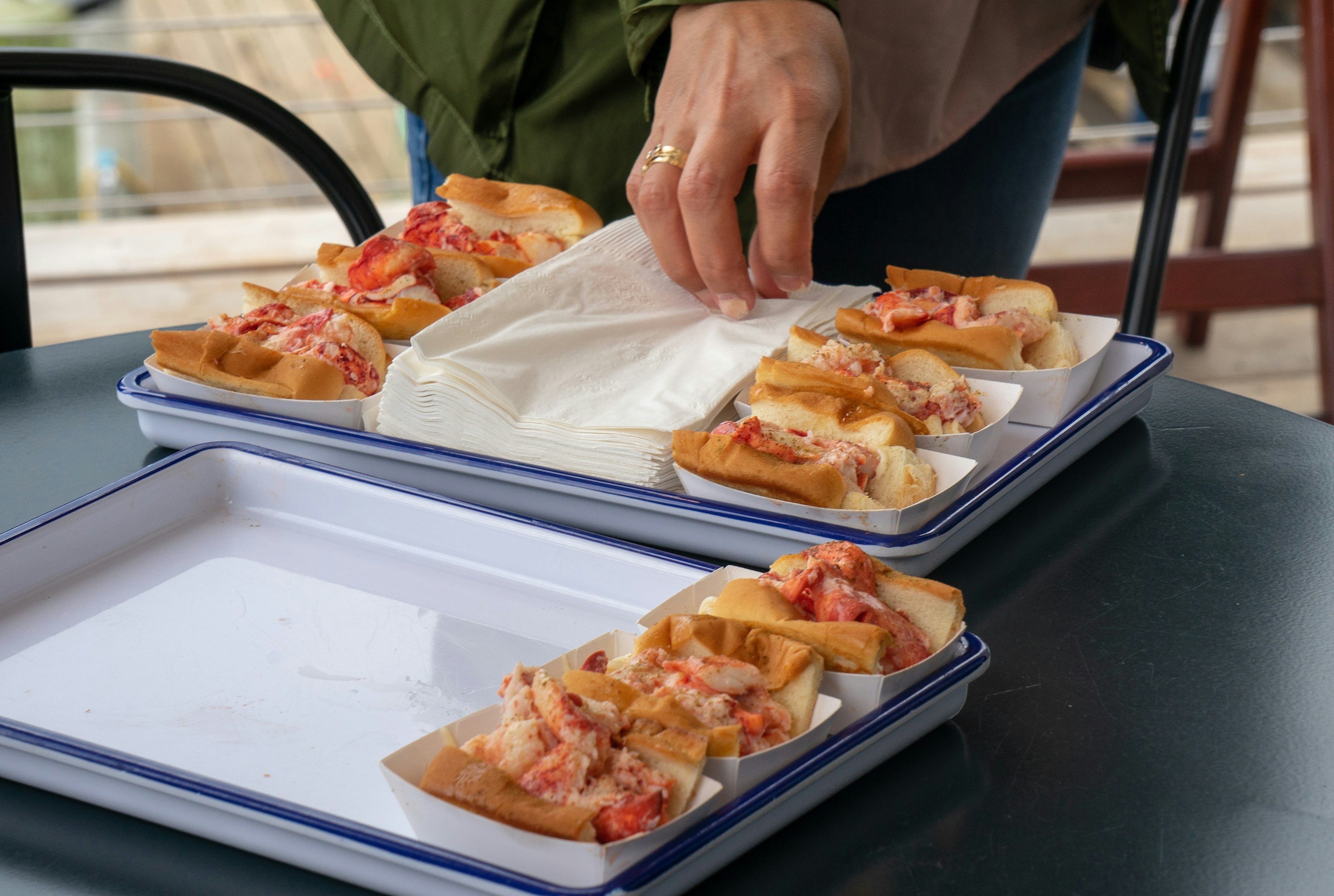 A hand wearing a gold wedding band reaches for a napkin from a pile set between two rows of Maine Lobster Rolls at Luke's in Portland. The rolls are in small paper trays and are heaped with bright red and pink lobster meat in white and blue enamel trays