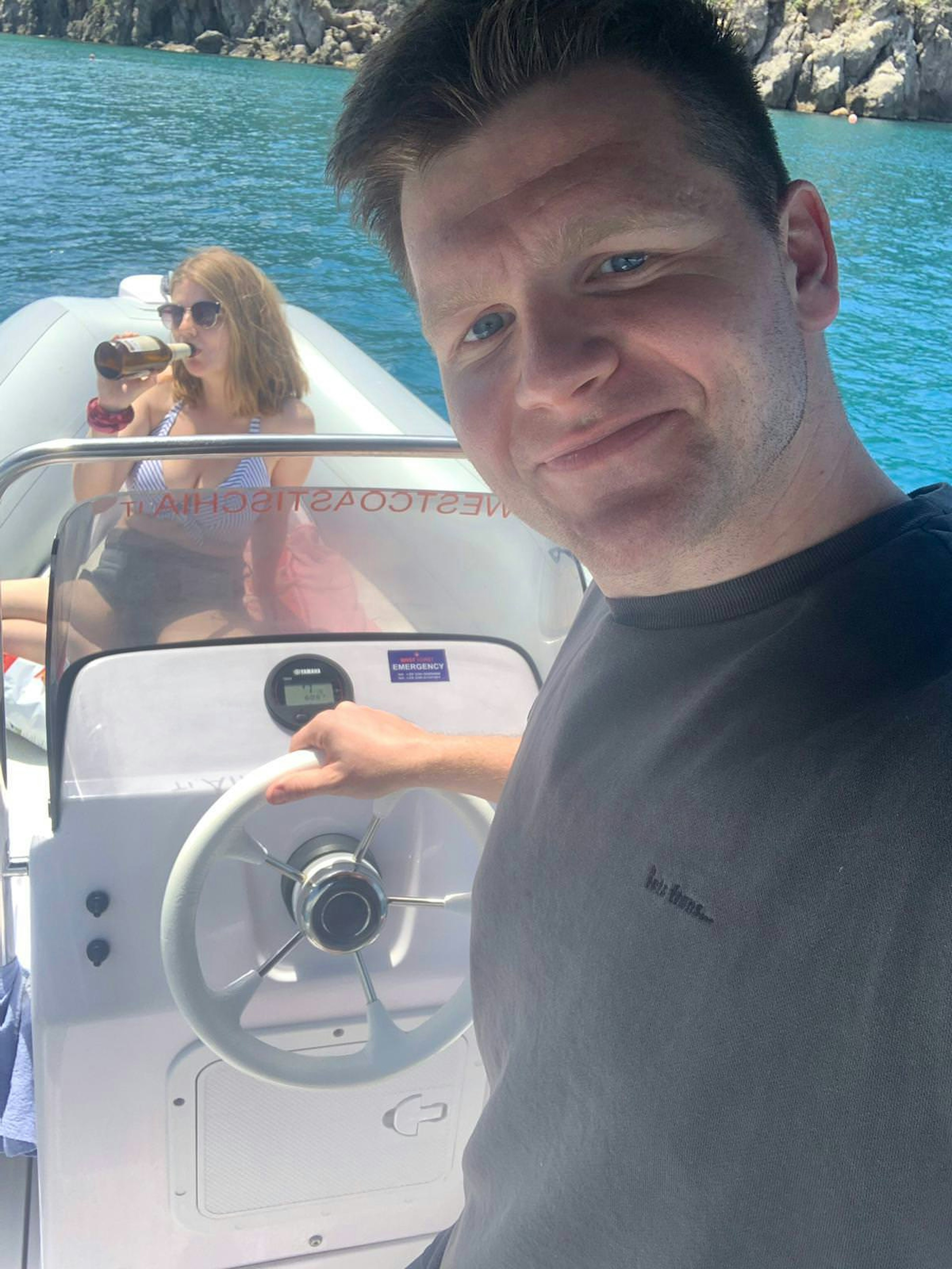 A man and a woman on a white speedboat at water. The man is taking a selfie while he holds the wheel, in the background the woman is swigging from a bottle of wine