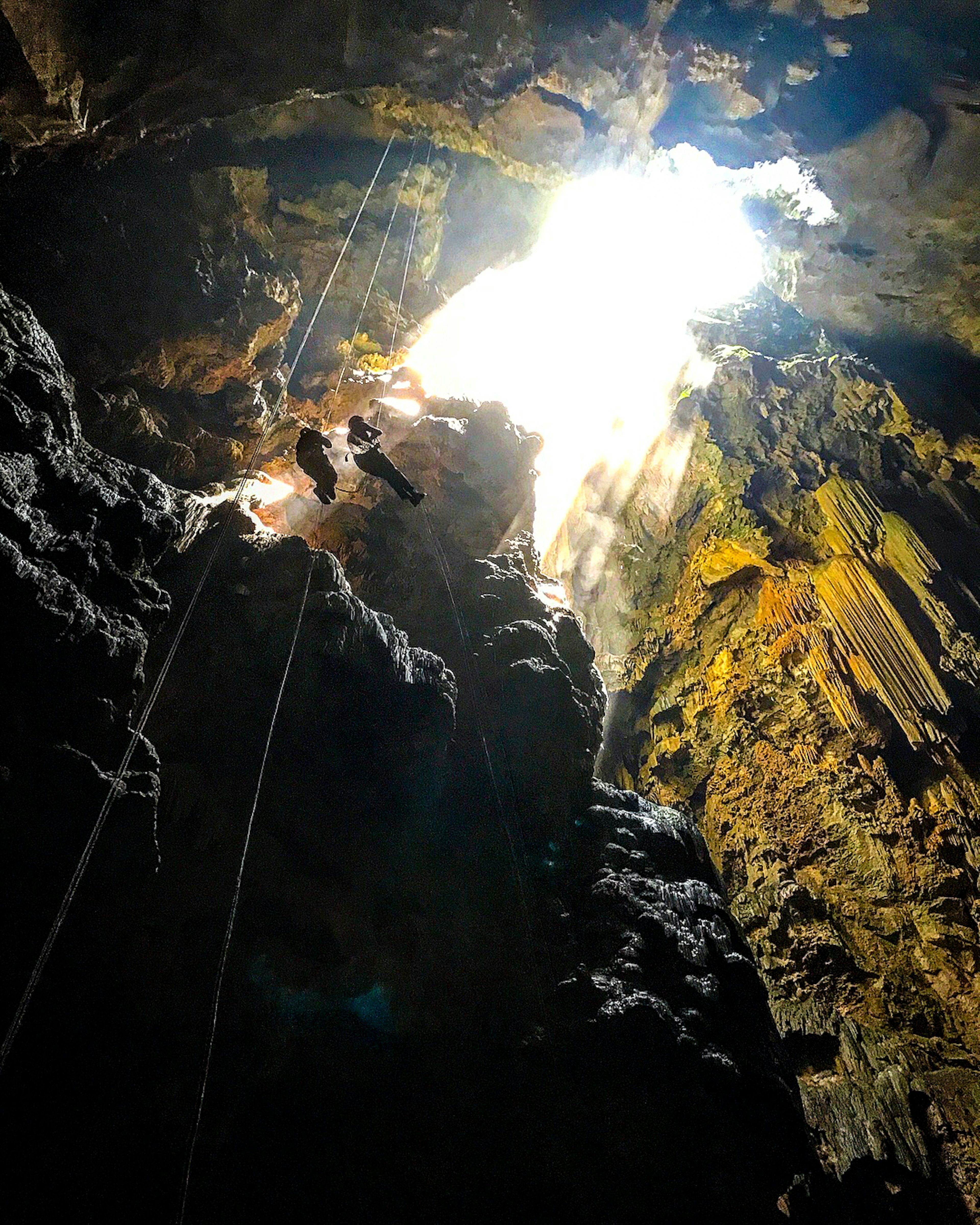 A shot from the floor of a tall cave looking up at its opening with two climbers hanging from ropes near the top