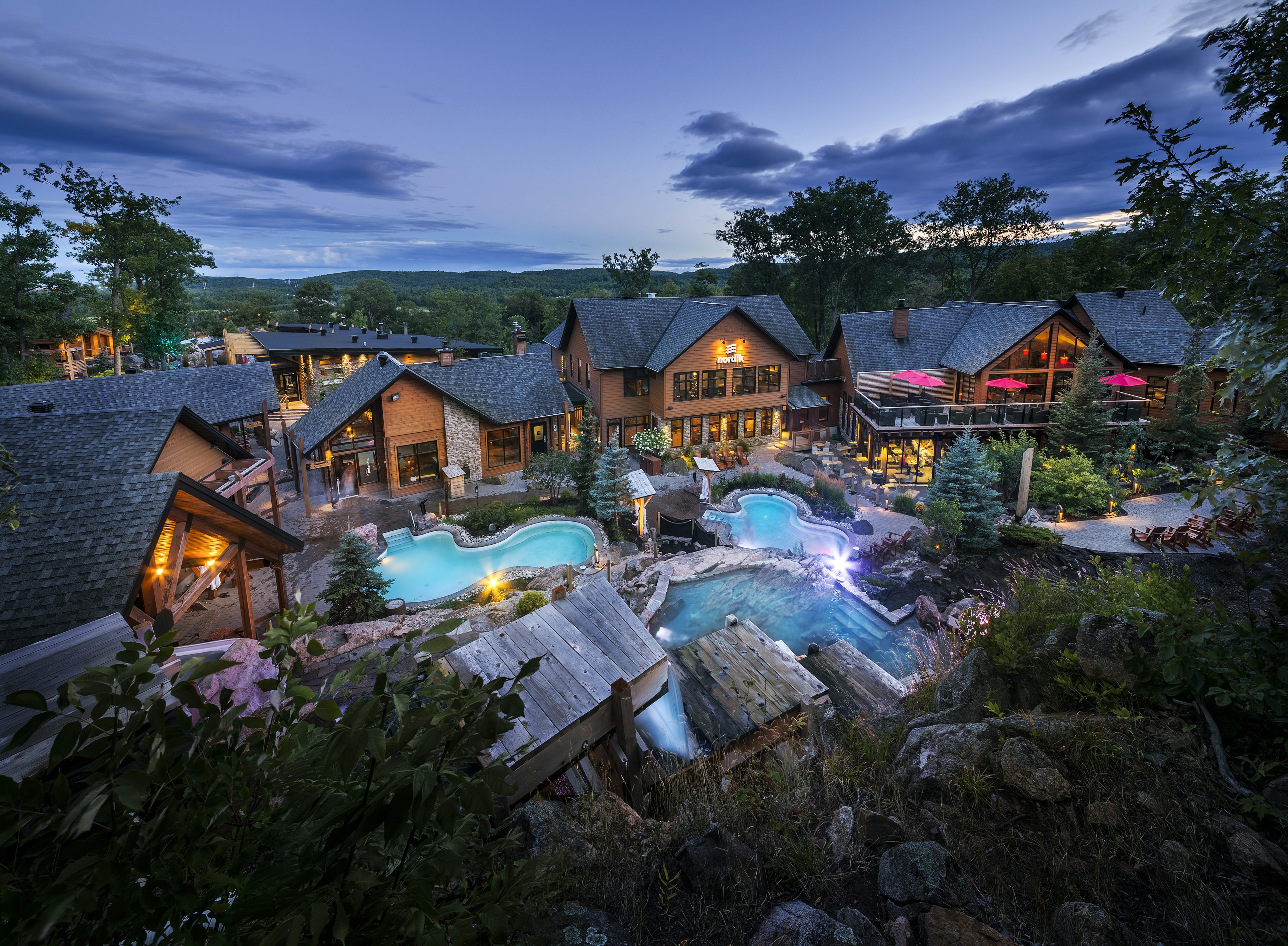 Aerial views of Nordik Spa Nature – brown houses circled around 3 pools