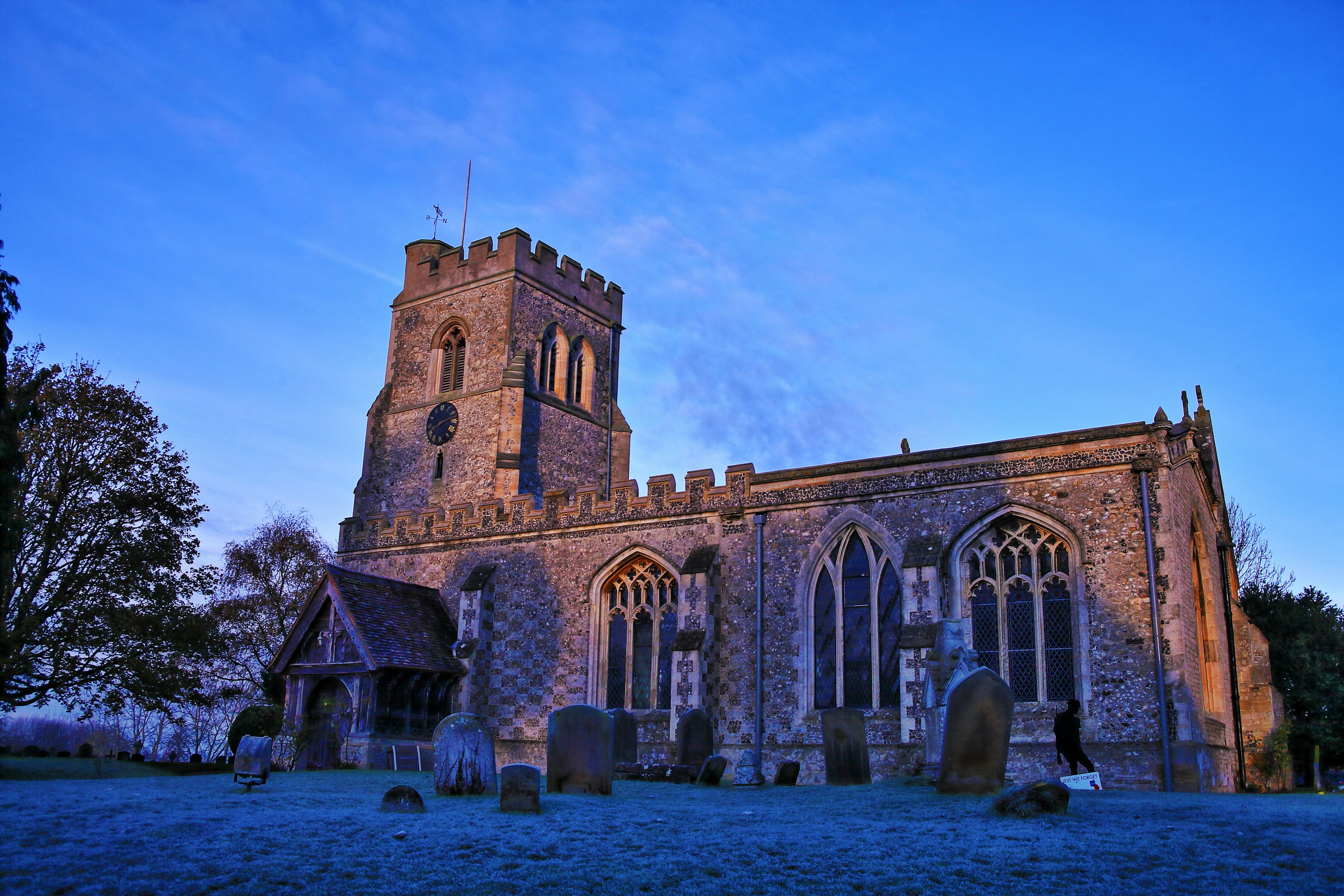 The outside of All Saints Church at dusk. A number of headstones are visible on the grass in front.