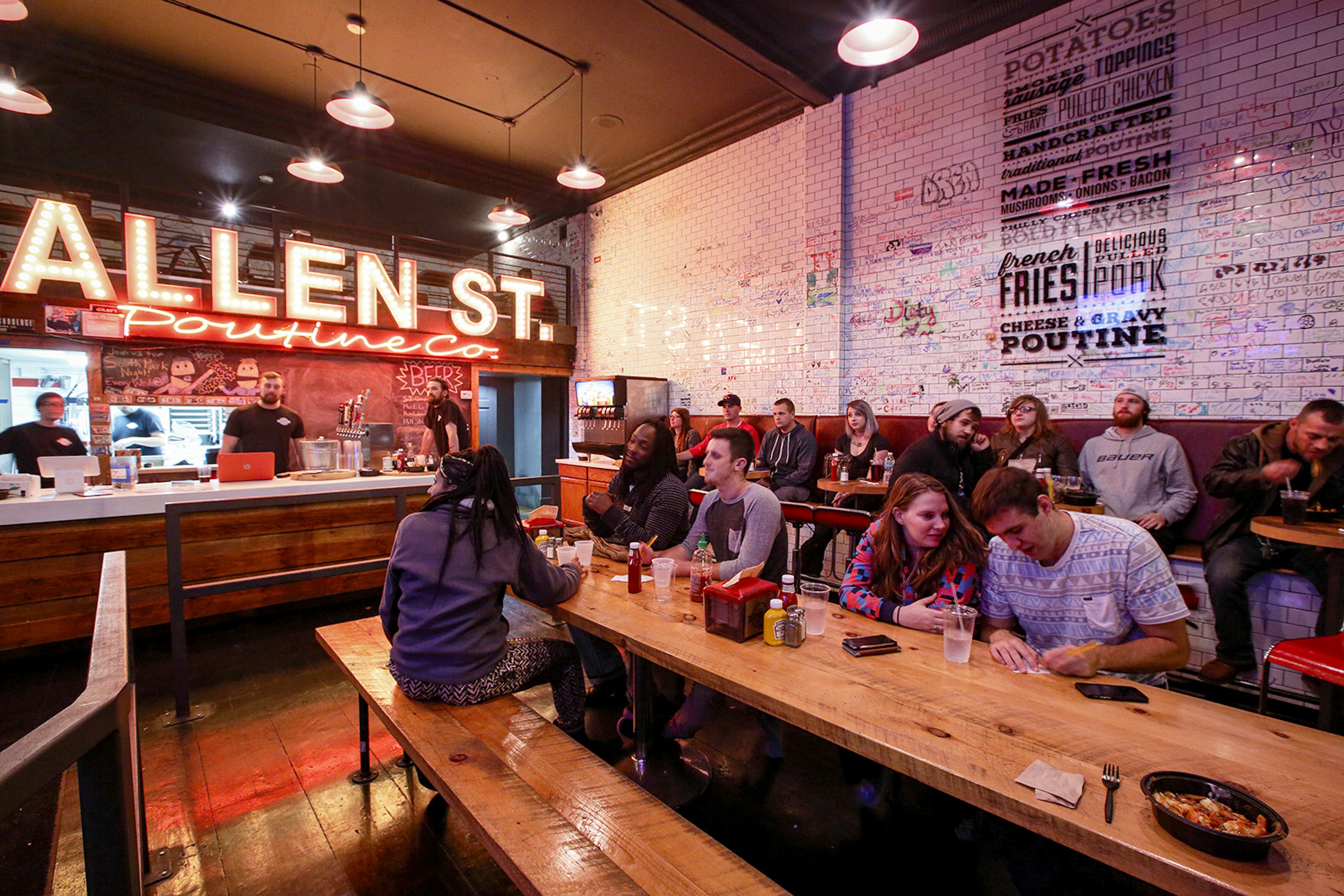 People sit at long wooden tables eating poutine. The counter is visible, with a large neon sign that says