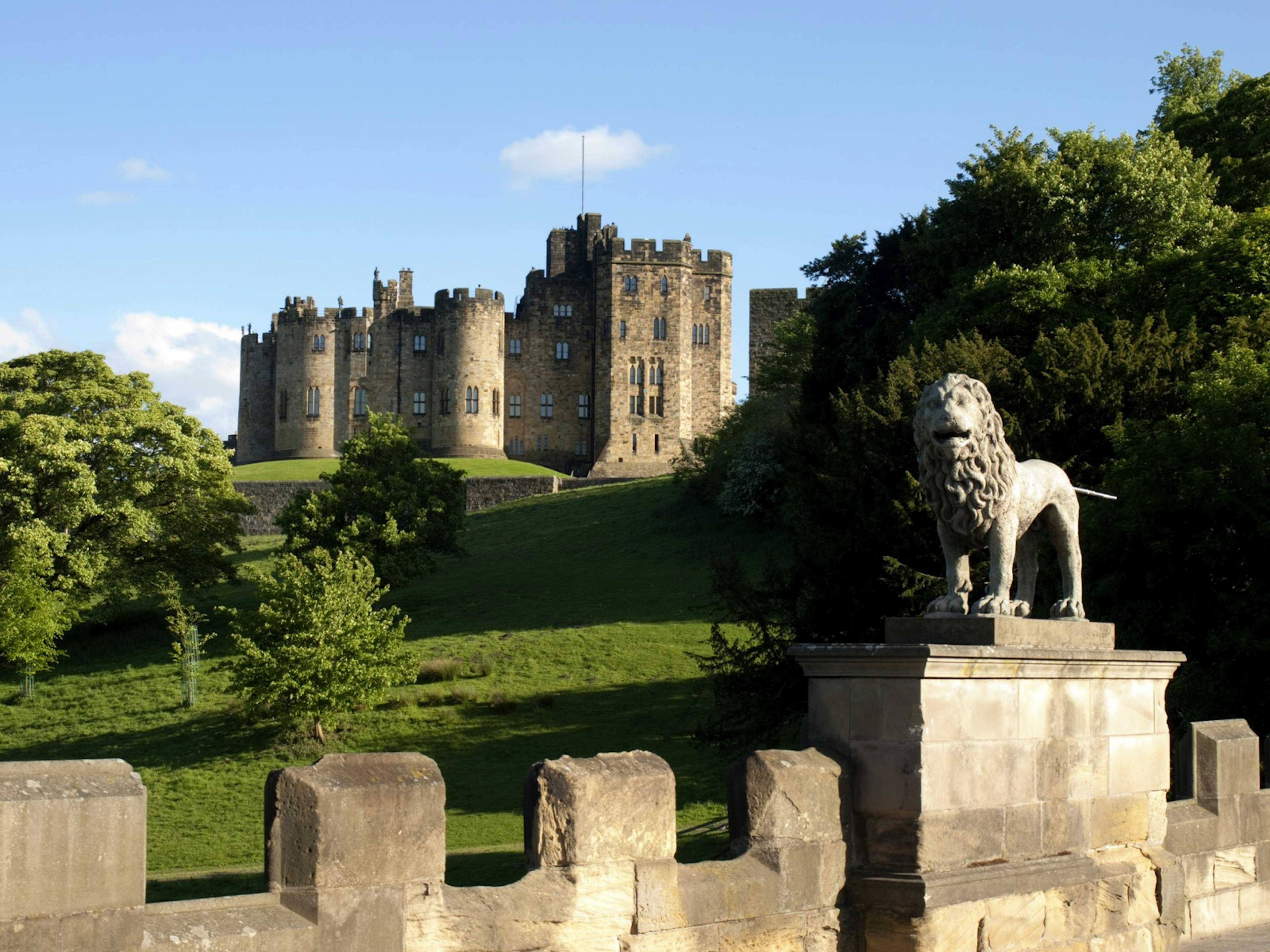 A castle sits on a grass covered hill with a lion and bridge in the foreground