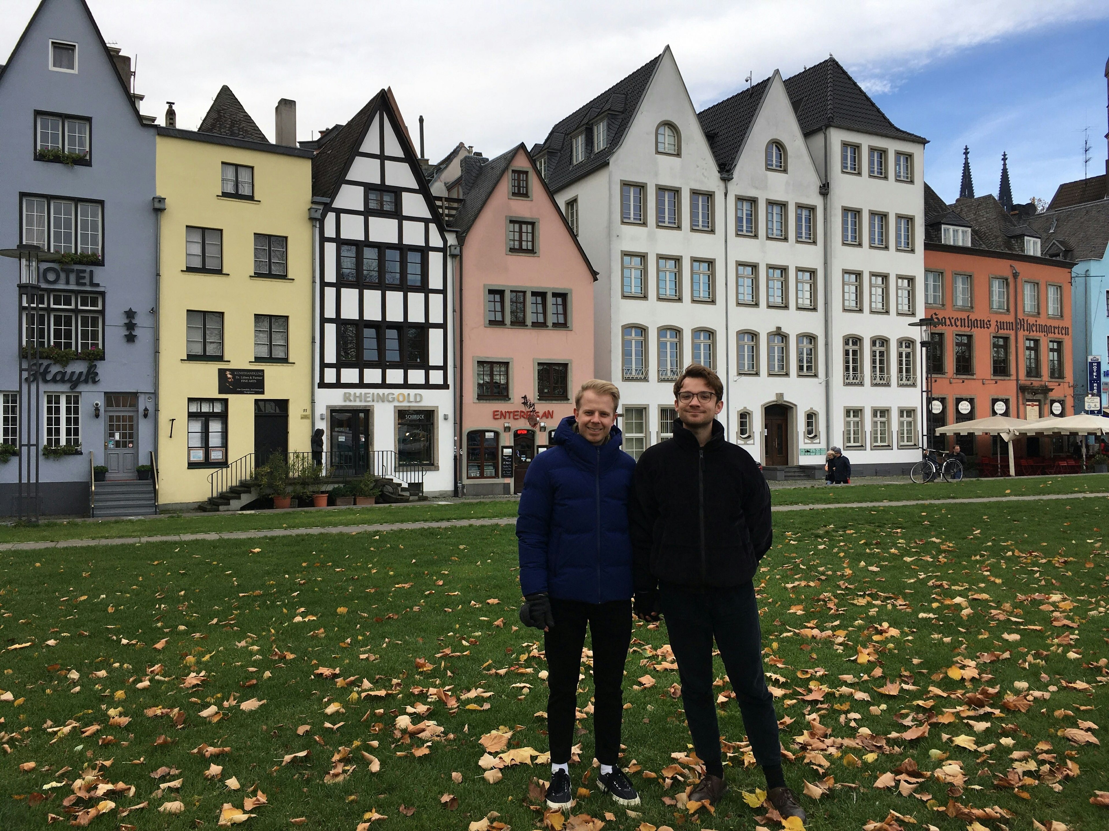 Two men stand on grass. In the background are typical narrow German buildings.