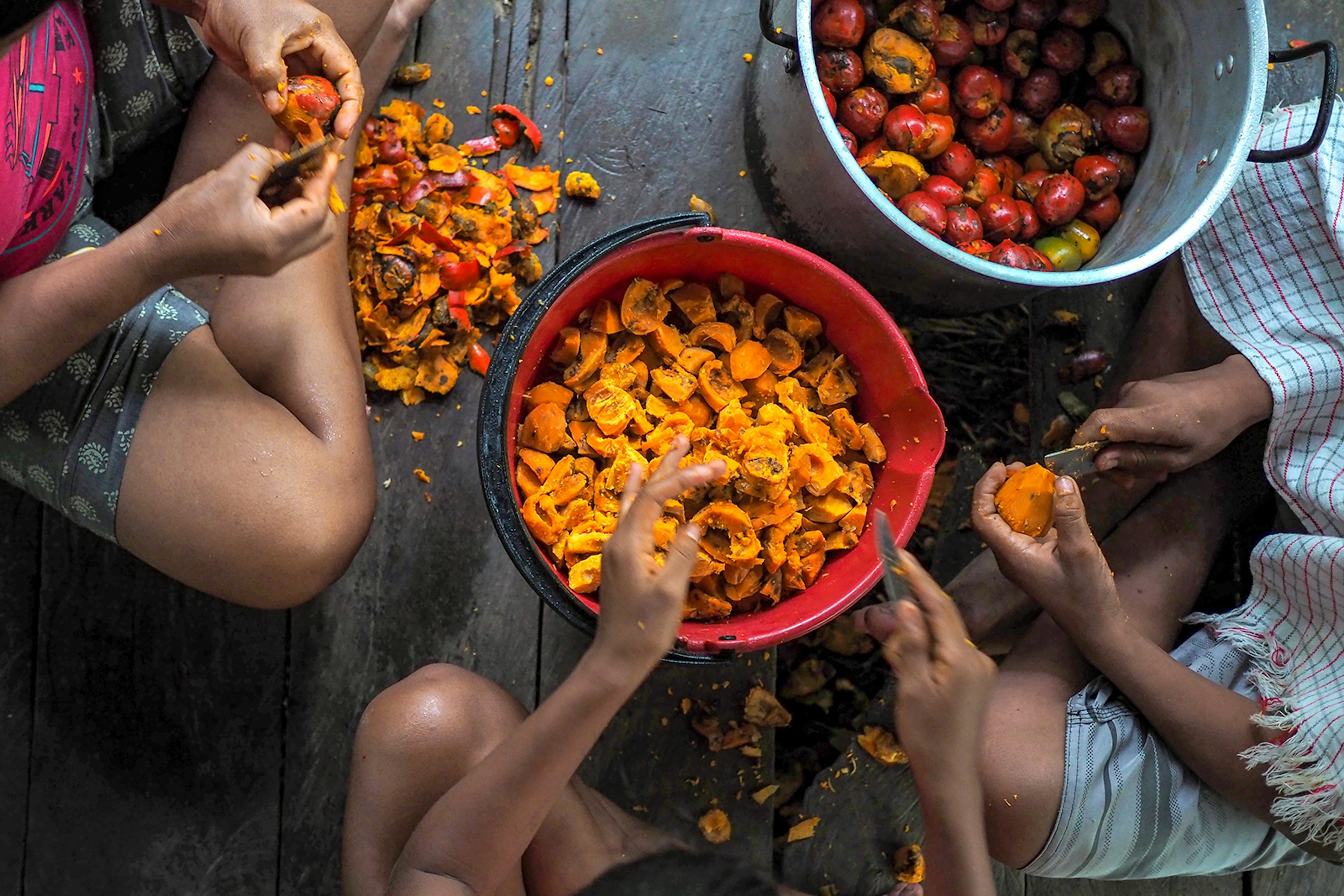 Three people sit around two buckets of orange fruit, peeling off the skins. Leticia, Colombia.