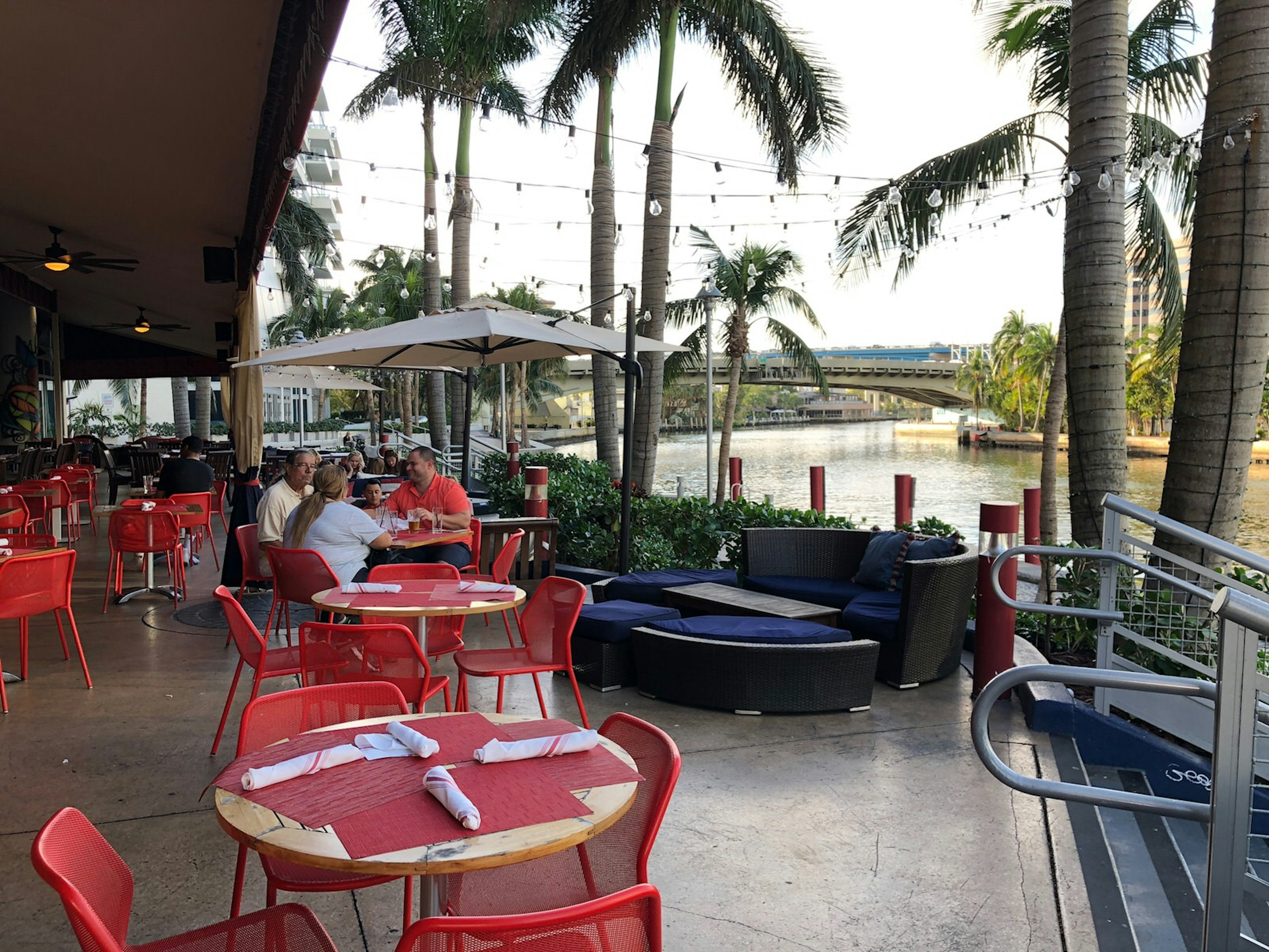 Red tablecloths lend a pop of color to a waterfront bar area with palm trees and sand beyond © Jackie Gutierrez-Jones