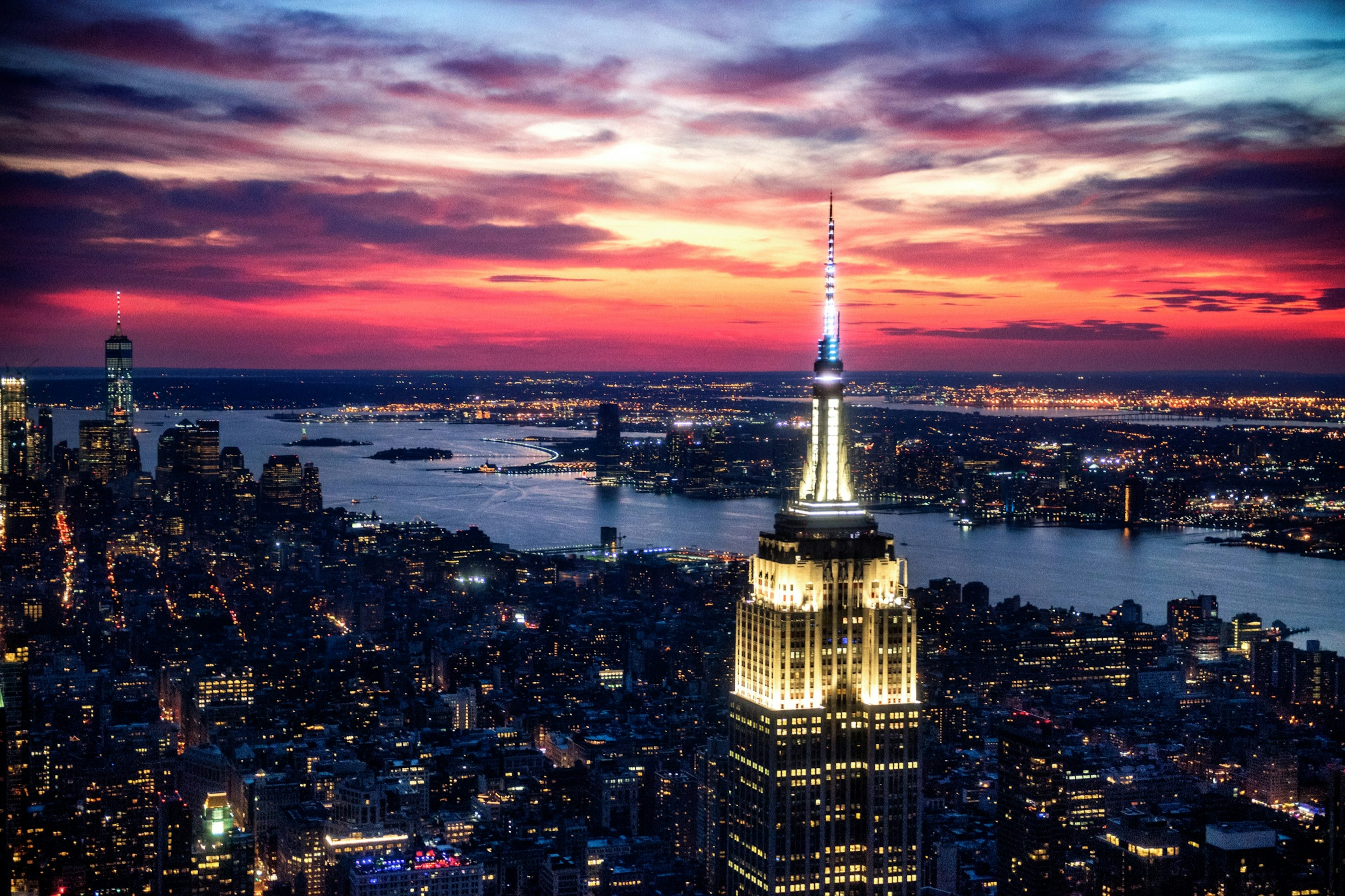 The top of the Empire State Building in New York City is illuminated at late dusk as the lights of the city are spread out below