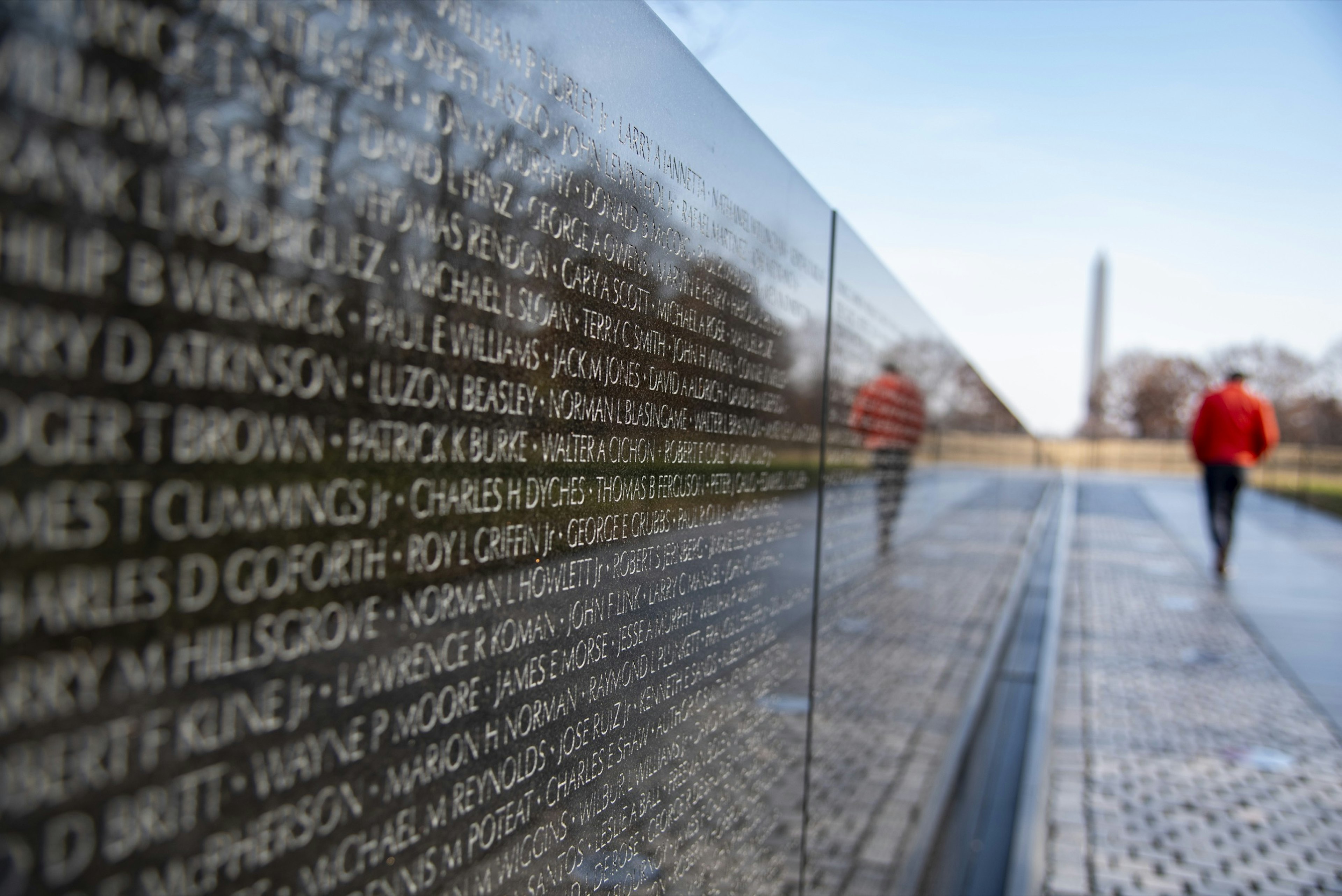 A man in a red coat is reflected in the black granite walls of the Vietnam Veterans Memorial as he walks toward the Washington Monument on a cold day