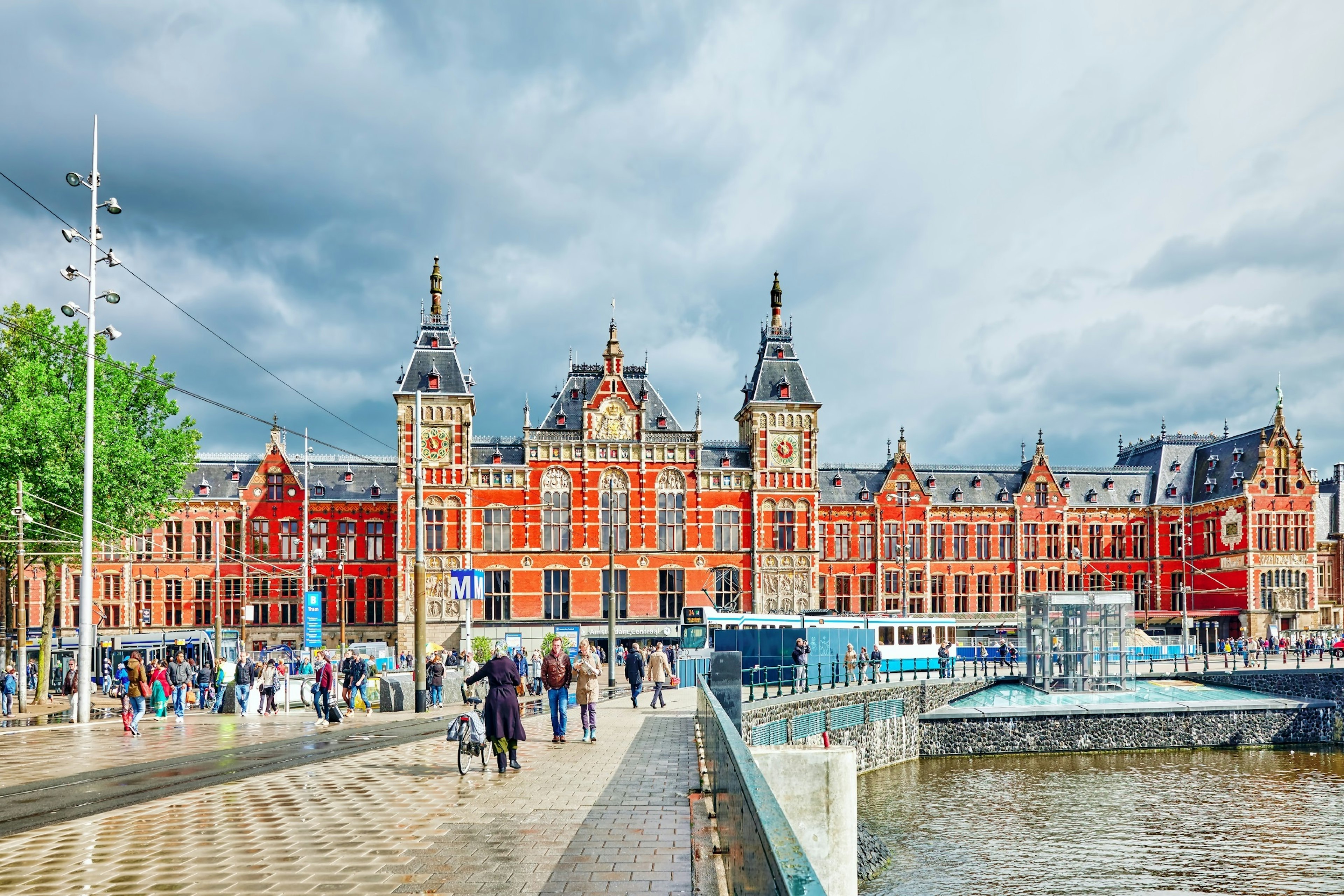 The exterior of a  large ornate red-brick building, with a canal and a pedestrianised section in the foreground