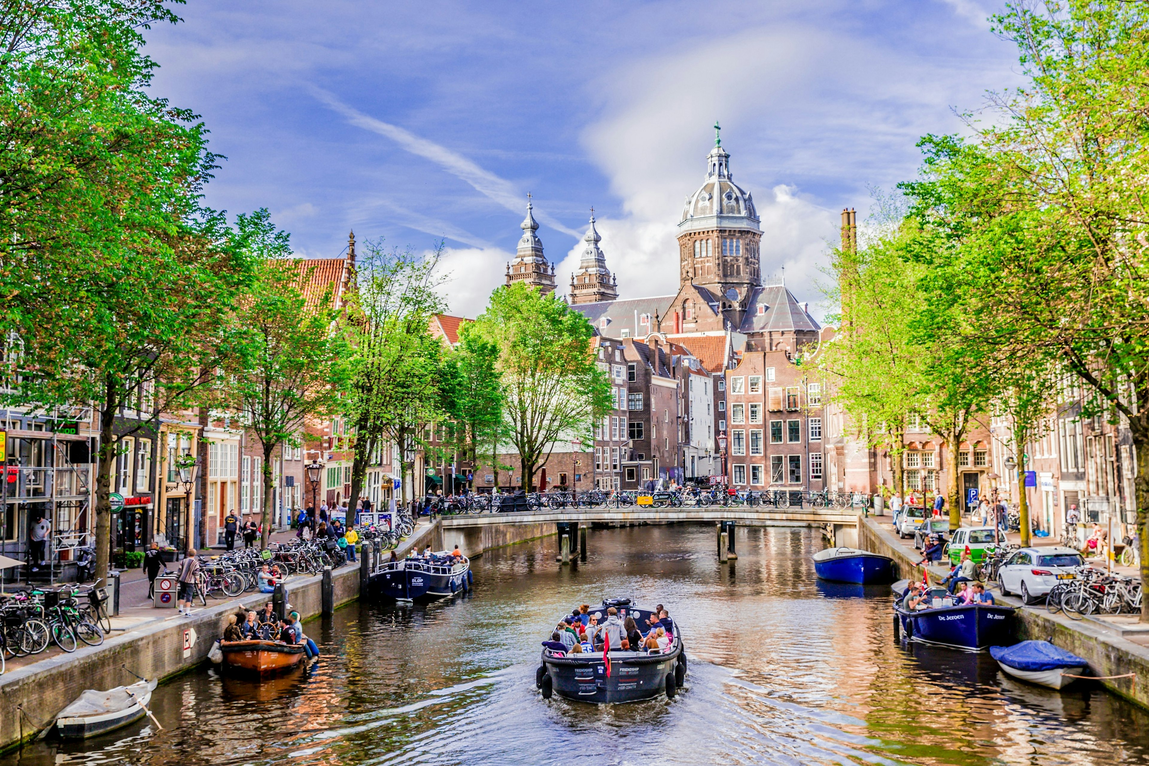 A canal boat travels along a canal. The canal is lined with parked bicycles, trees and red-brick buildings