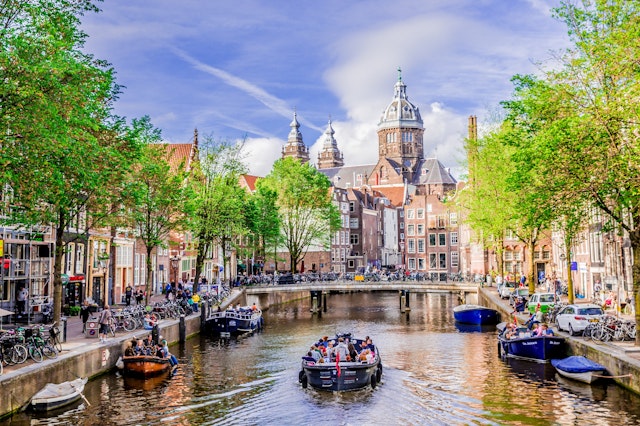 A canal boat travels along a canal. The canal is lined with parked bicycles, trees and red-brick buildings