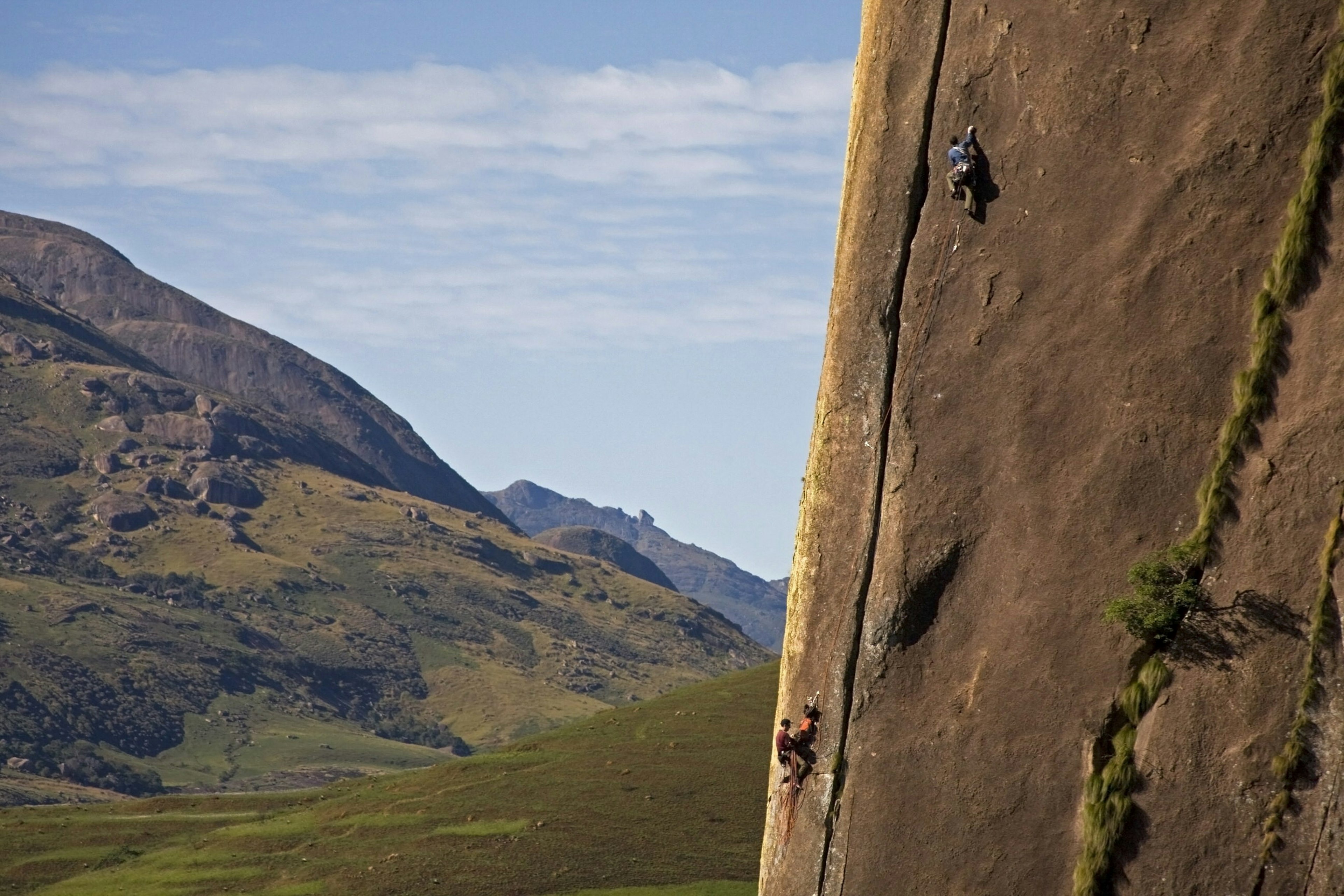 Two rock climbers making their way up a sheer rock face with the left half of the images being dominated by grass-covered hills. Southern Madagascar, Africa