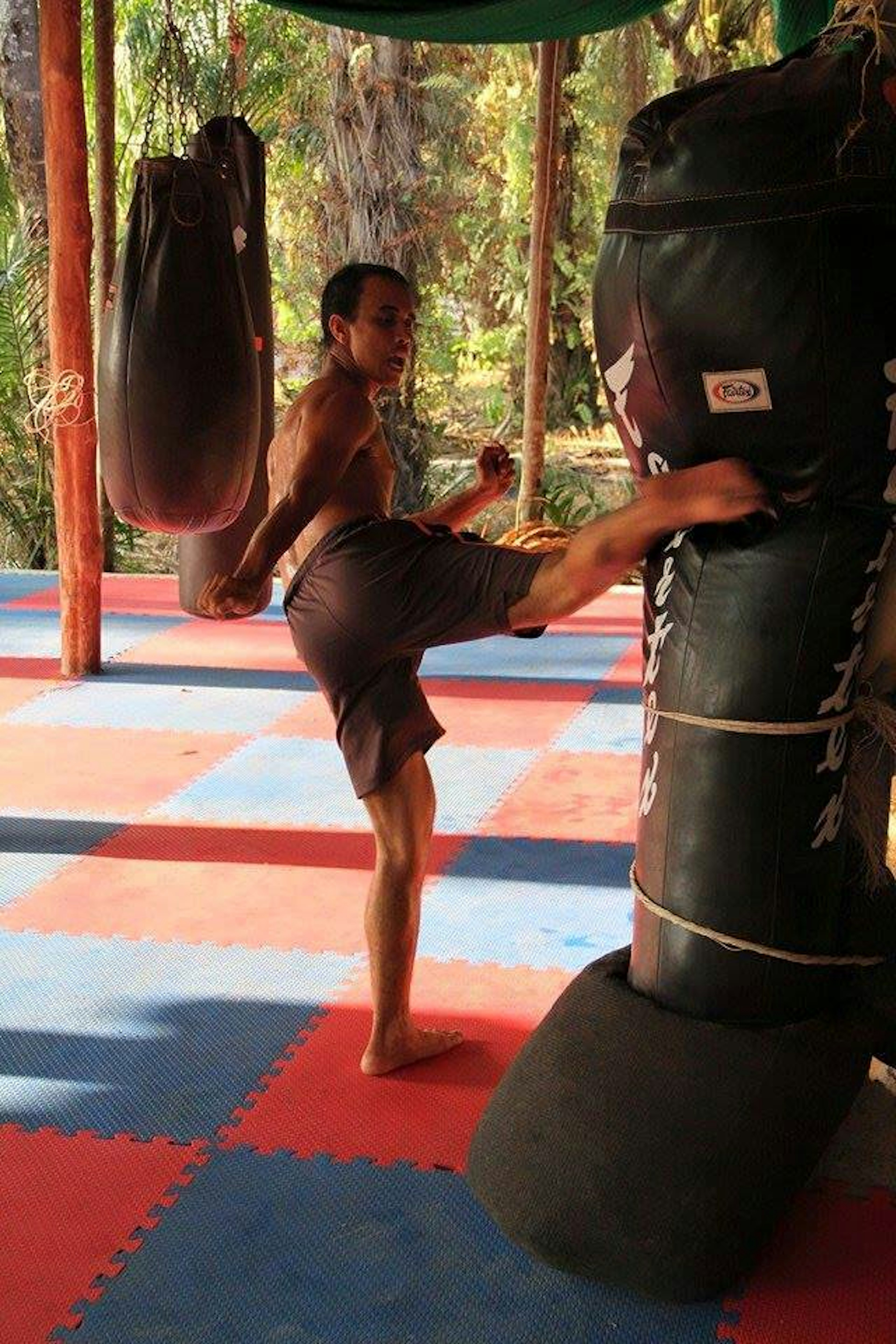 A man kicks a punchbag in an outdoor gym. The floor is chequered blue and red tiles.