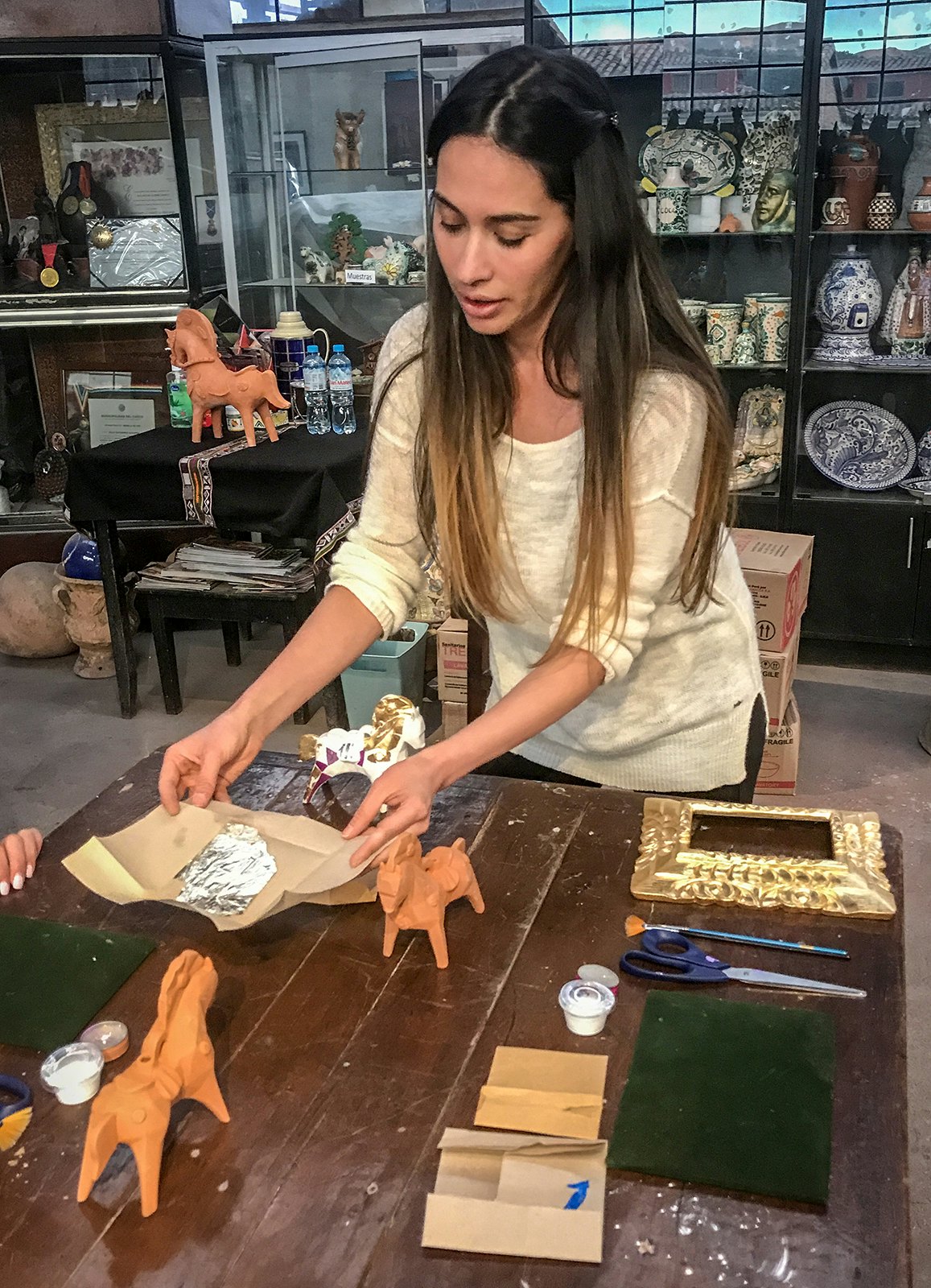 A woman leans forward holding a sheet of foil, with two ceramic horses sitting on the table in front of her