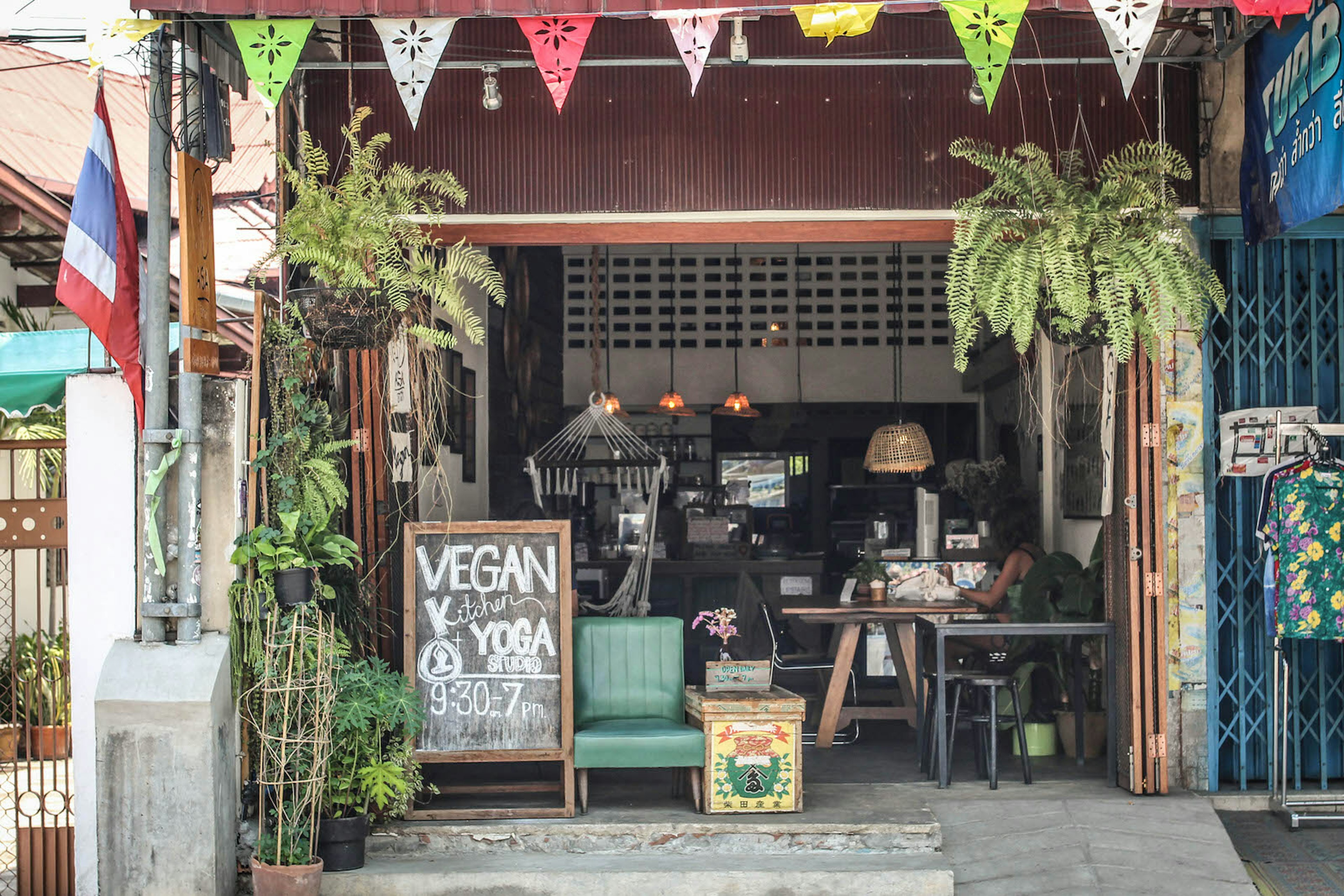 The exterior of Asa cafe in Chinag Mai. There is colourful bunting and hanging plants hung in front of the cafe and a blackboard reading 'VEGAN Kitchen + YOGA STUDIO' with class times underneath. There's comfy seating and a cafe counter visible inside.